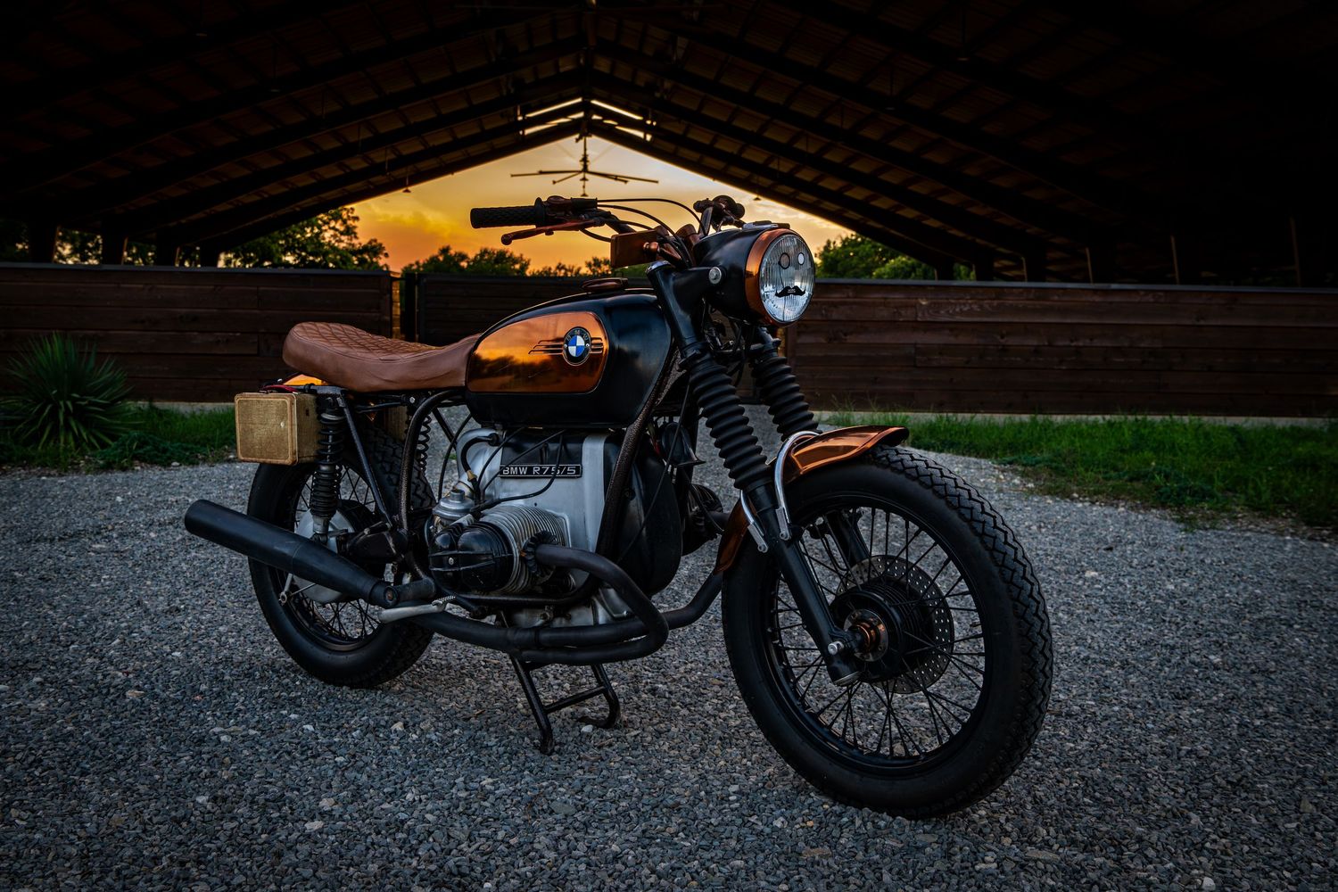Vintage BMW motorcycle with brown leather seat parked on gravel at sunset under a wooden pavilion.