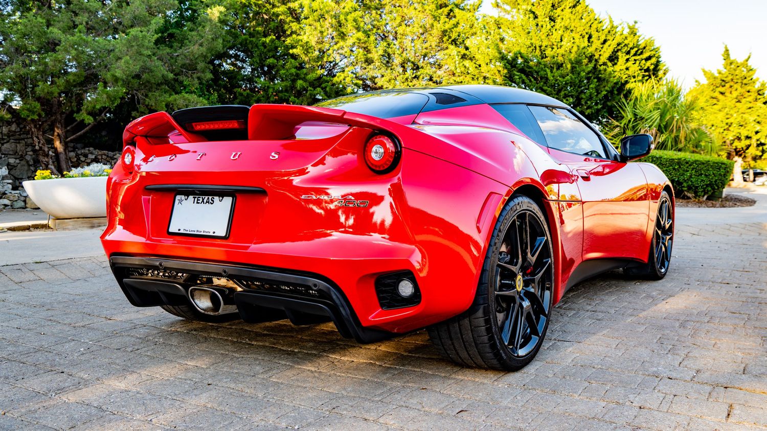 Red Lotus Evora sports car rear view with black alloy wheels and spoiler parked on a driveway surrounded by trees.