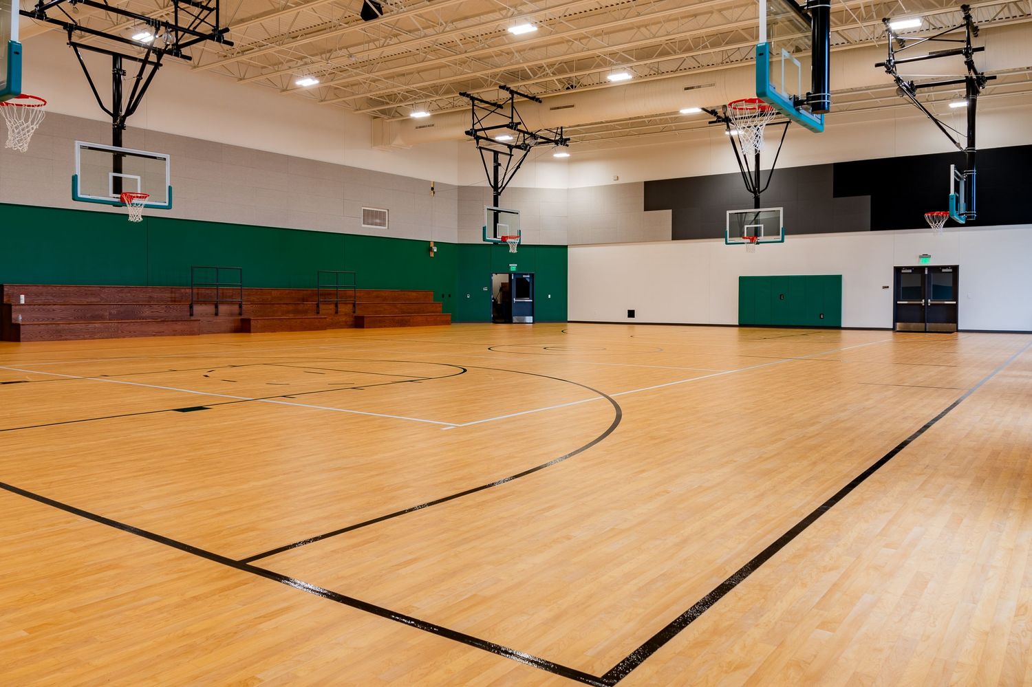 Indoor basketball court with wooden flooring and multiple hoops visible in a well-lit gymnasium space.