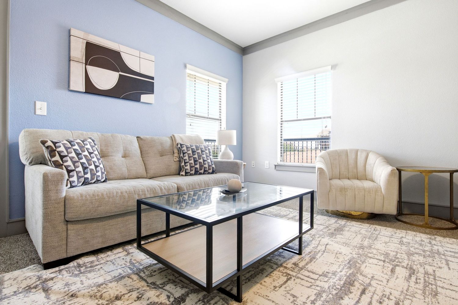 Modern living room with beige tufted sofa, glass coffee table, accent chair, and blue walls with natural light.