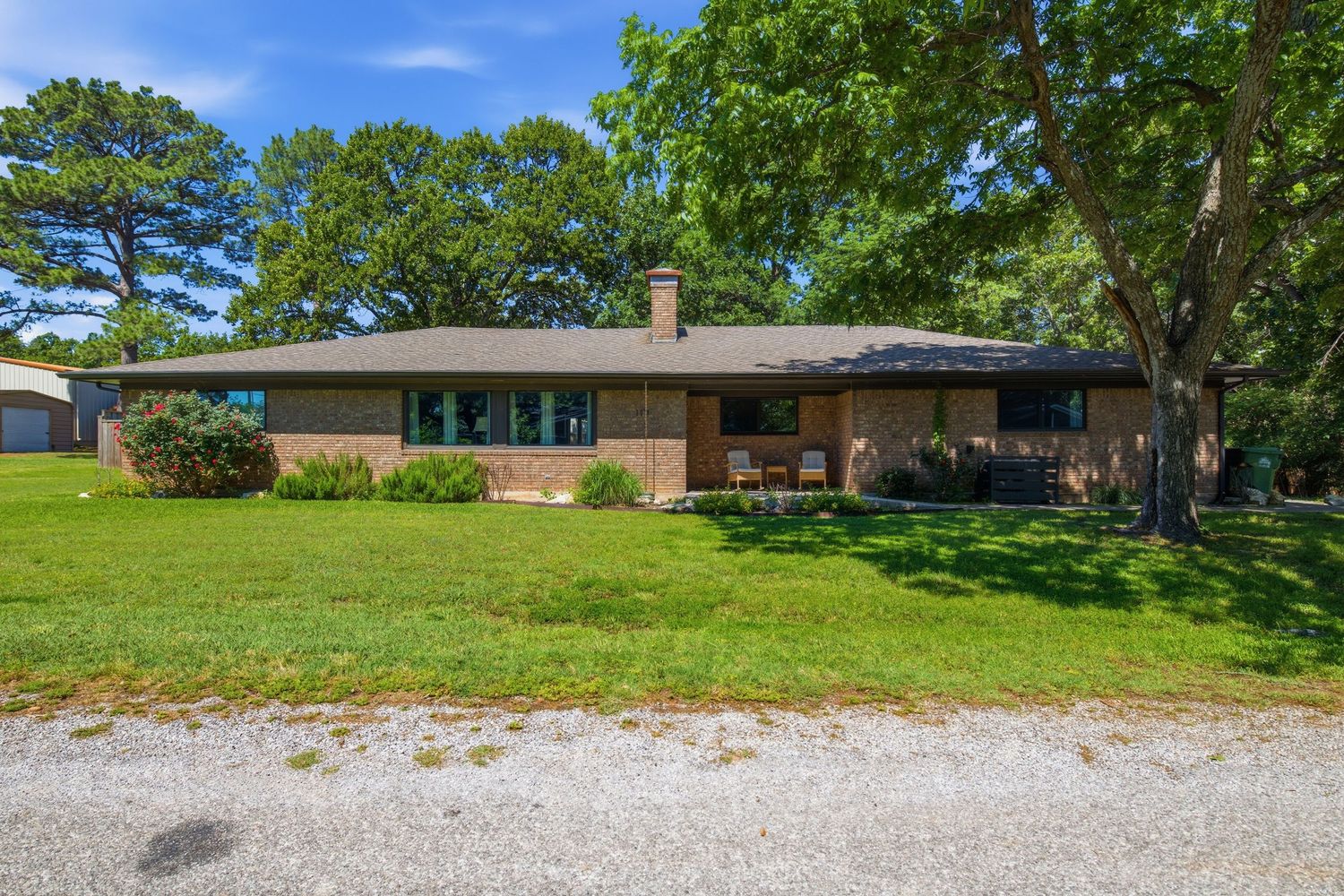 Brick ranch-style home with large green lawn, mature shade trees, and gravel driveway on a sunny day.