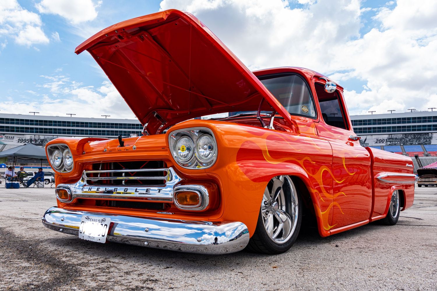 Custom orange lowrider 1958 Chevy Apache pickup truck with flames, chrome details, and hood open at a car show.