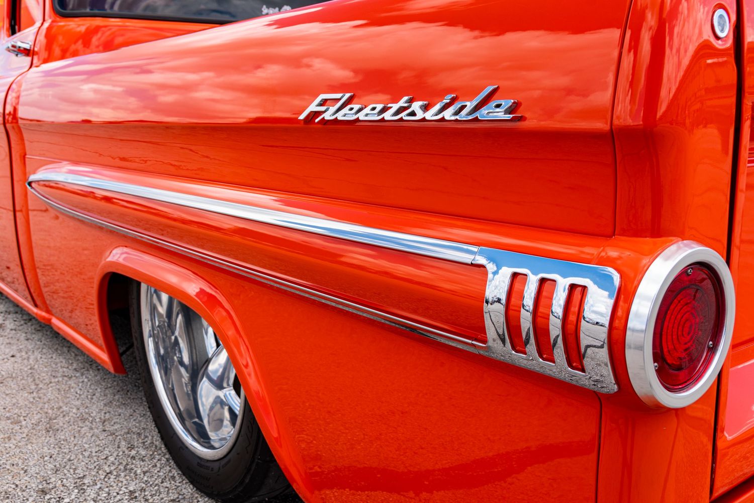 Vintage orange Chevrolet Fleetside pickup truck detail showing chrome badge and tail fin at a classic car show.