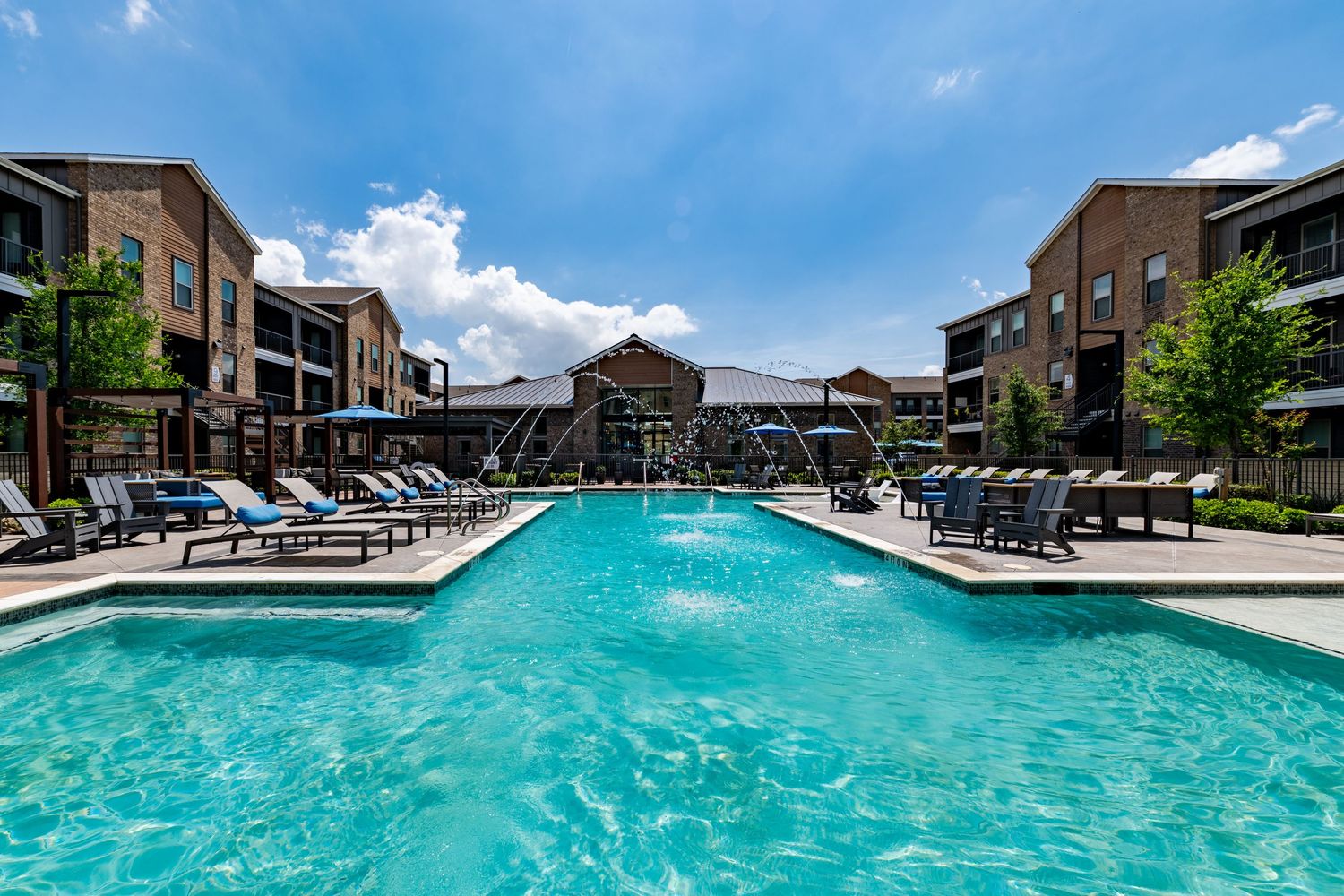 Luxury apartment complex pool with lounge chairs, blue water, and brick buildings under a bright summer sky.
