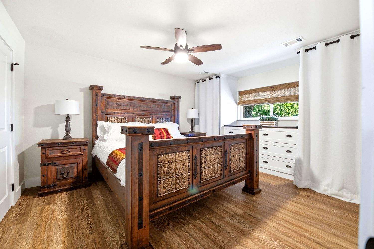 Rustic wooden four-poster bed with carved details in a bright bedroom featuring hardwood floors and a ceiling fan.