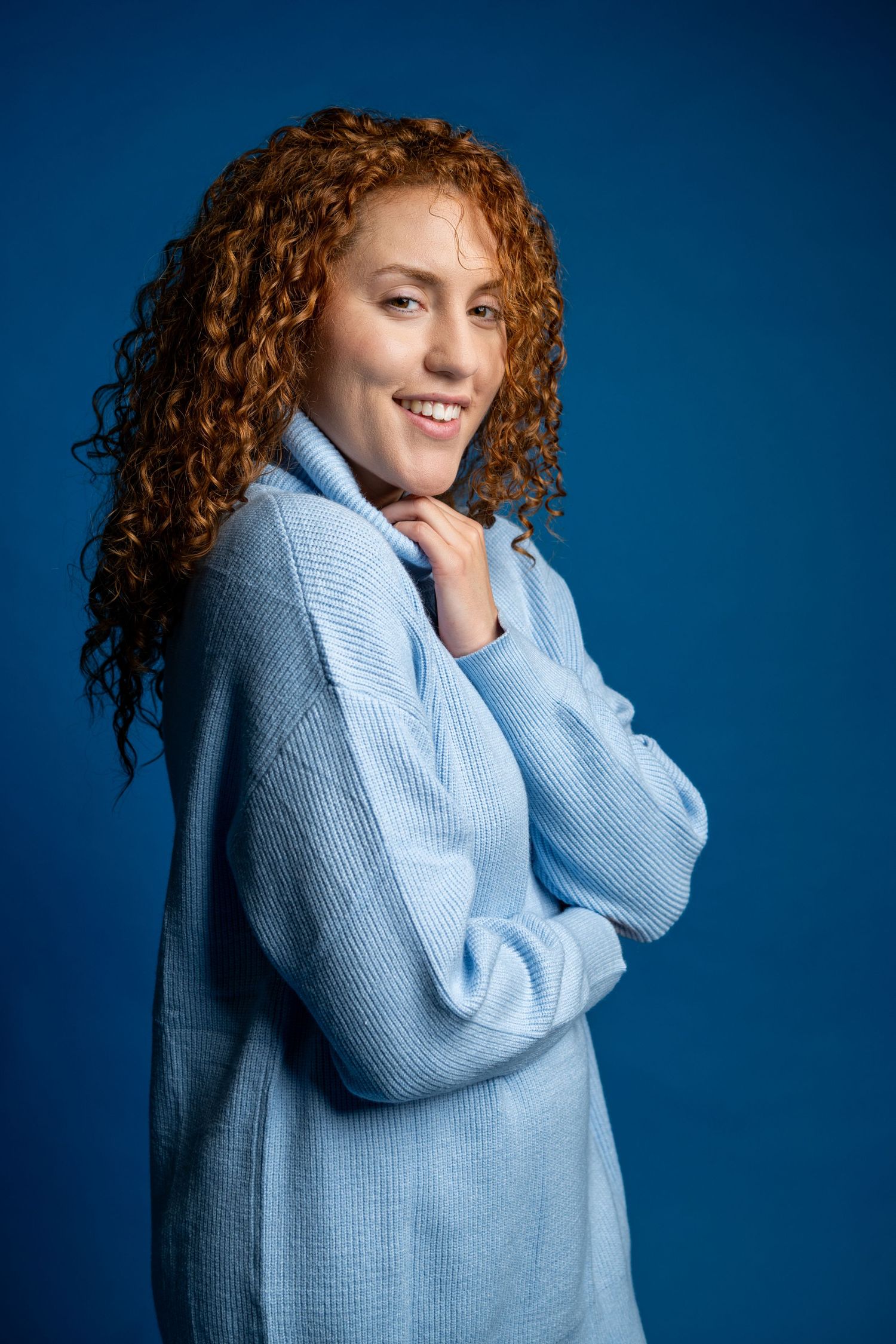 Smiling woman with curly red hair wearing a light blue turtleneck sweater posed against a blue background.