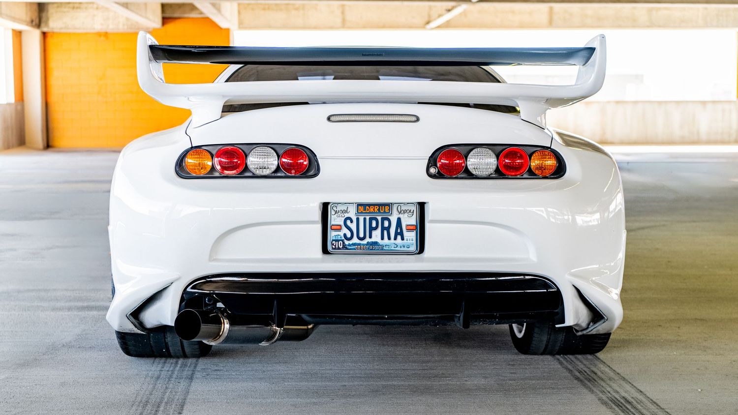 Rear view of a white Toyota Supra with a large spoiler, custom license plate reading SUPRA, and dual exhaust tips.