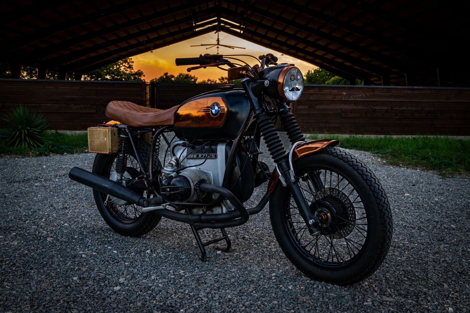 Vintage BMW motorcycle with brown leather seat parked on gravel at sunset near a wooden shelter.