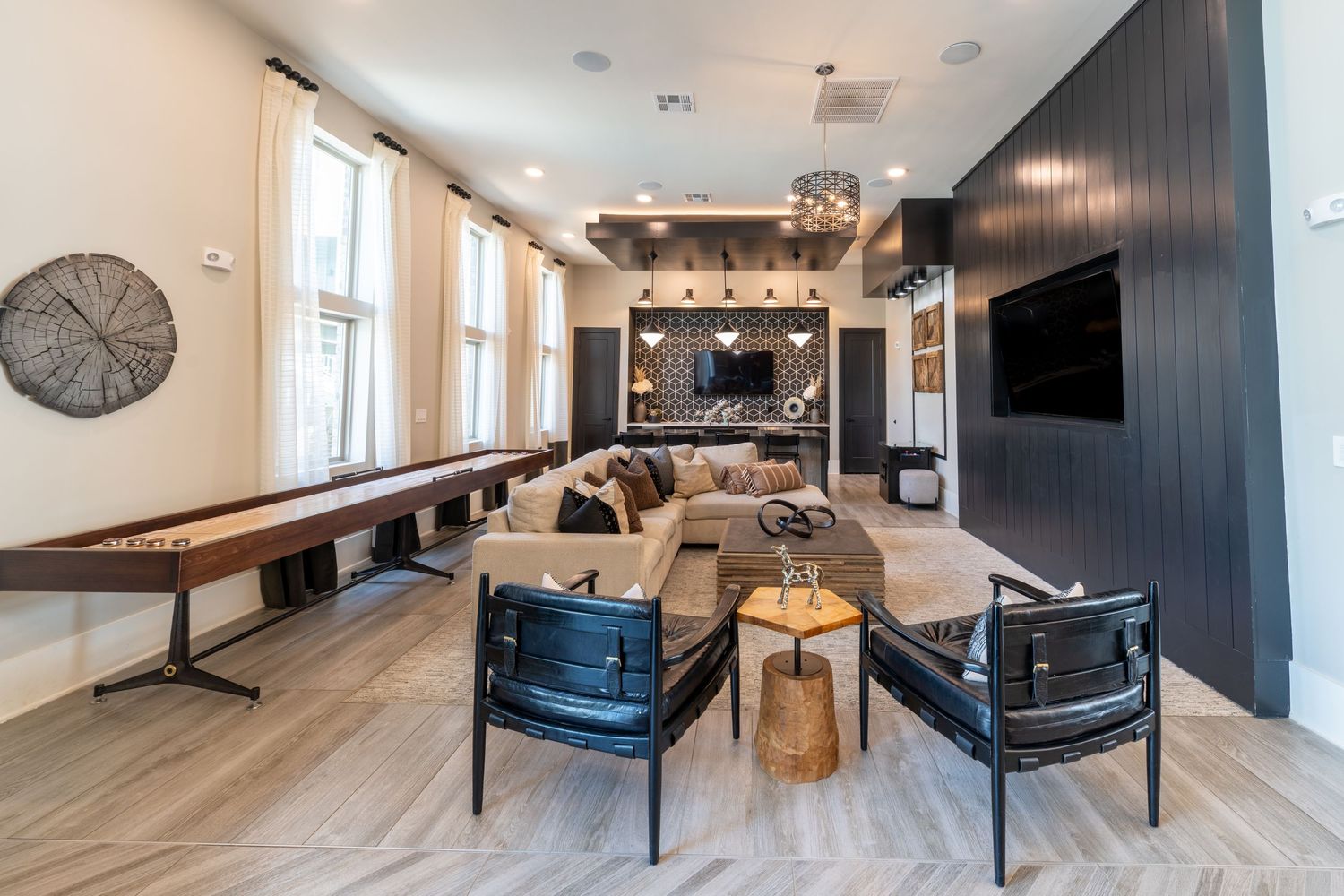 Modern apartment clubroom with shuffleboard table, sectional sofa, black accent wall, mounted TV, and stylish seating on light wood floors.