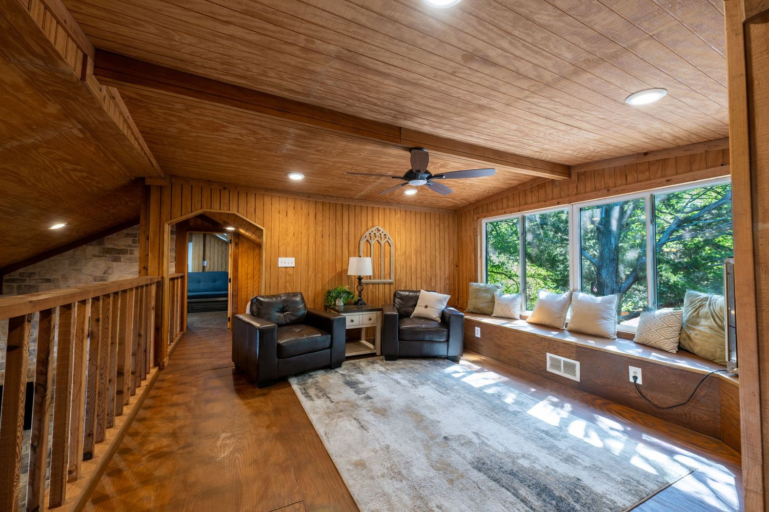 Cozy wood-paneled living room with leather chairs, built-in window seat, ceiling fan, and large windows overlooking lush greenery.