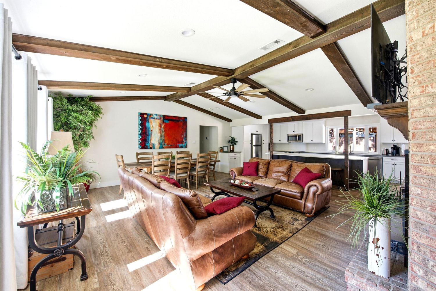 Spacious living room with vaulted wood beam ceilings, leather sofas, hardwood floors, and an open dining area with lush greenery.