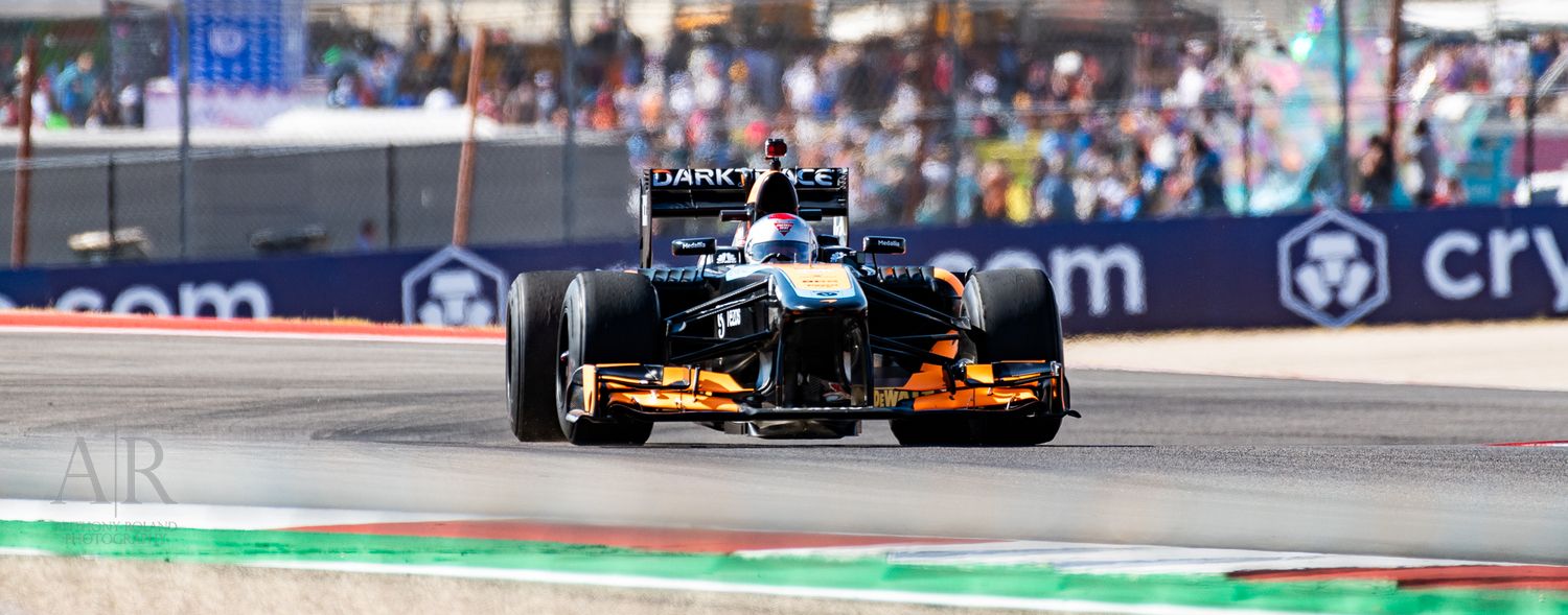 Formula 1 race car navigates turn on circuit with crowd visible in background.