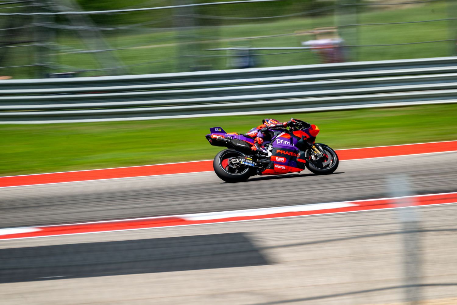 Racing motorcycle speeds along a racetrack curve with red and white curbing on a sunny day.