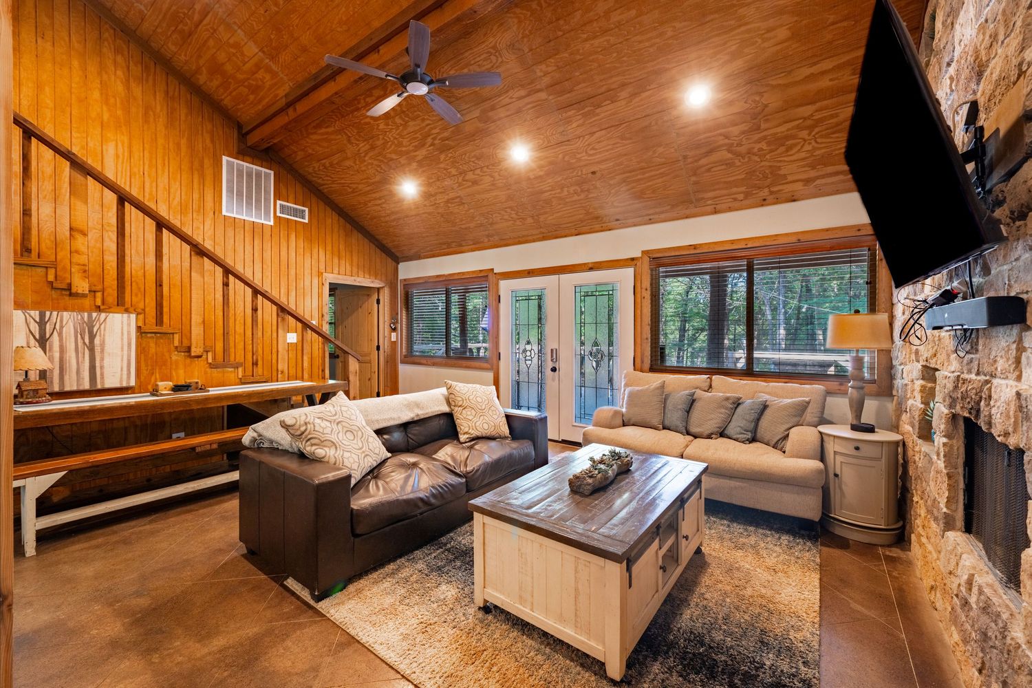 Cozy cabin living room with vaulted wood ceiling, stone fireplace, sectional sofa, and French doors leading to wooded views.
