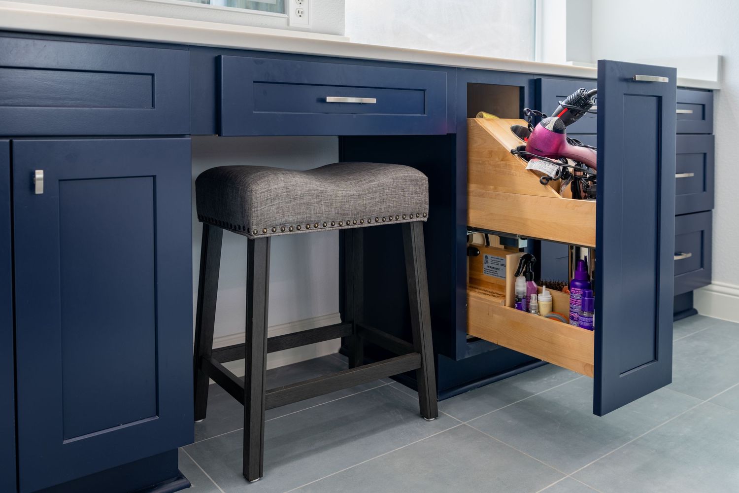 Navy blue kitchen island with pull-out storage drawer, Dyson vacuum organizer, and upholstered gray bar stool on tile floor.