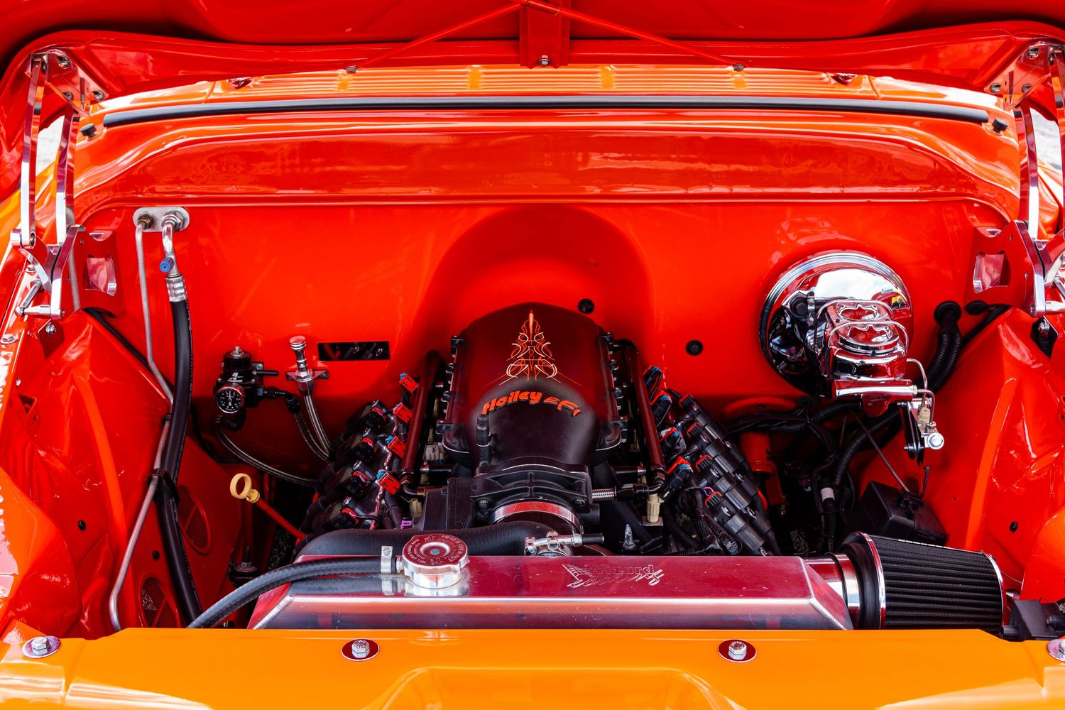 Bright orange classic car engine bay with chrome supercharger and custom red valve covers on display.