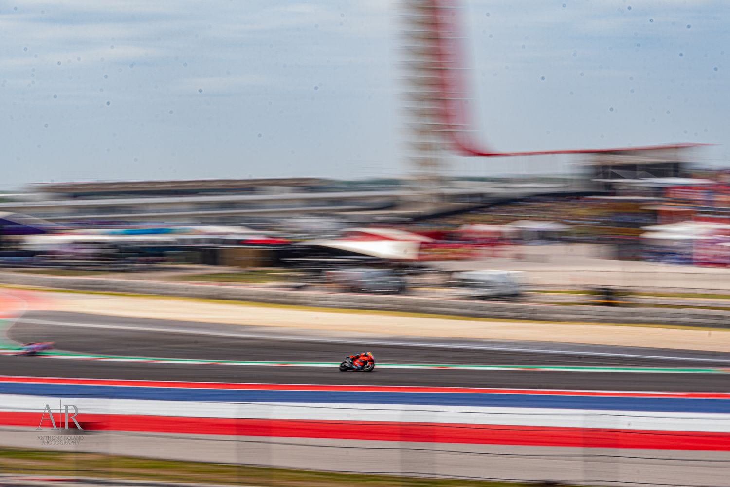 Blurred motorcycle speeds past on race track with control tower in background.