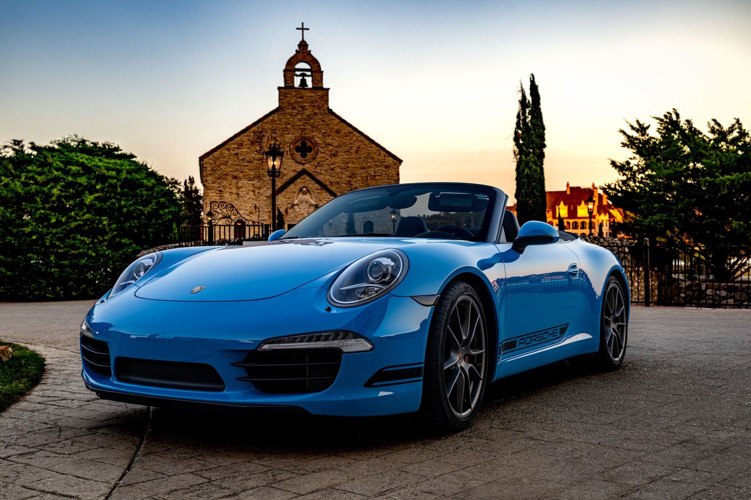 A vibrant blue Porsche 911 convertible parked on a stone plaza at sunset with a historic church in the background.