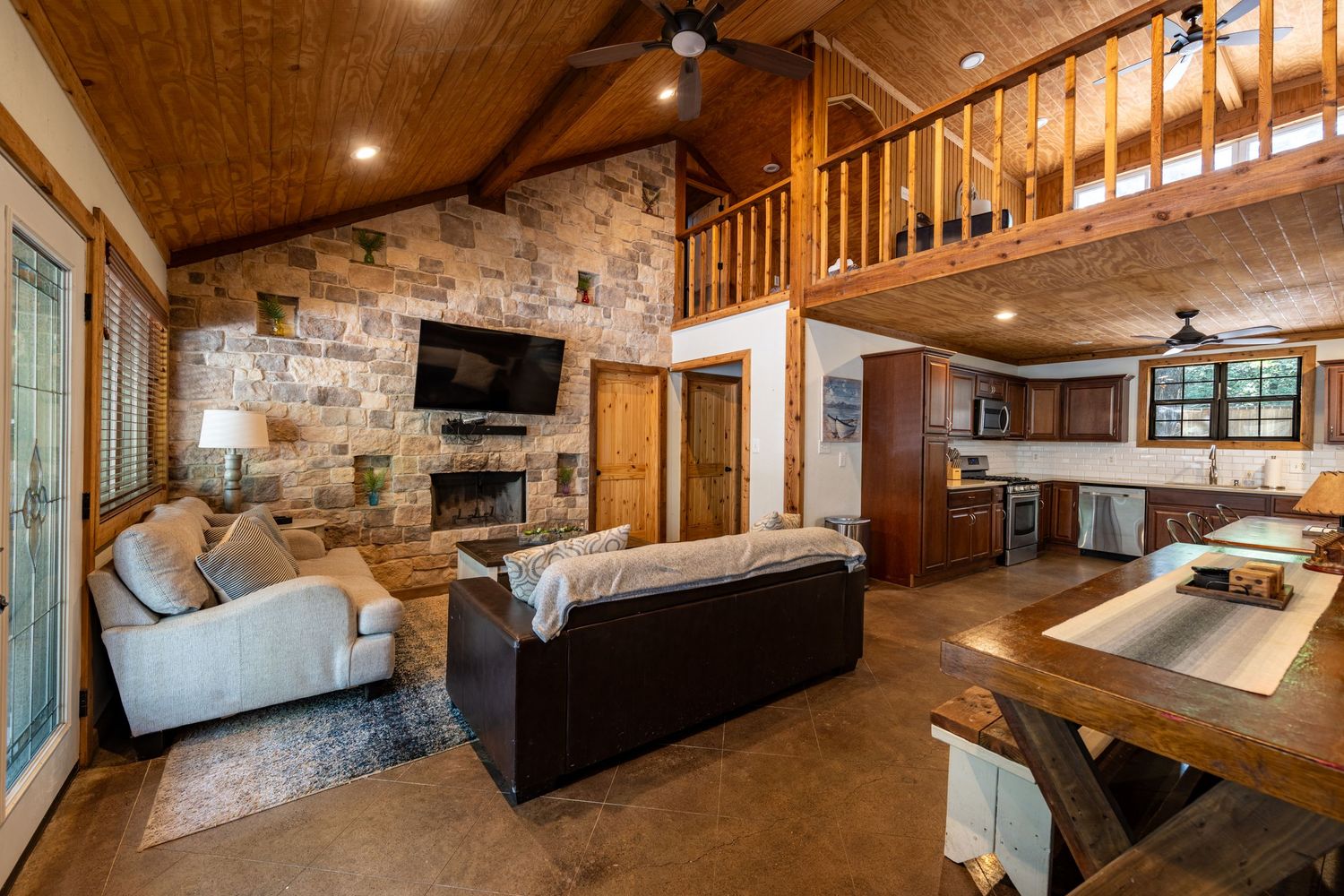 Rustic cabin living room with stone fireplace, vaulted wood ceiling, loft, open kitchen, and cozy seating area.