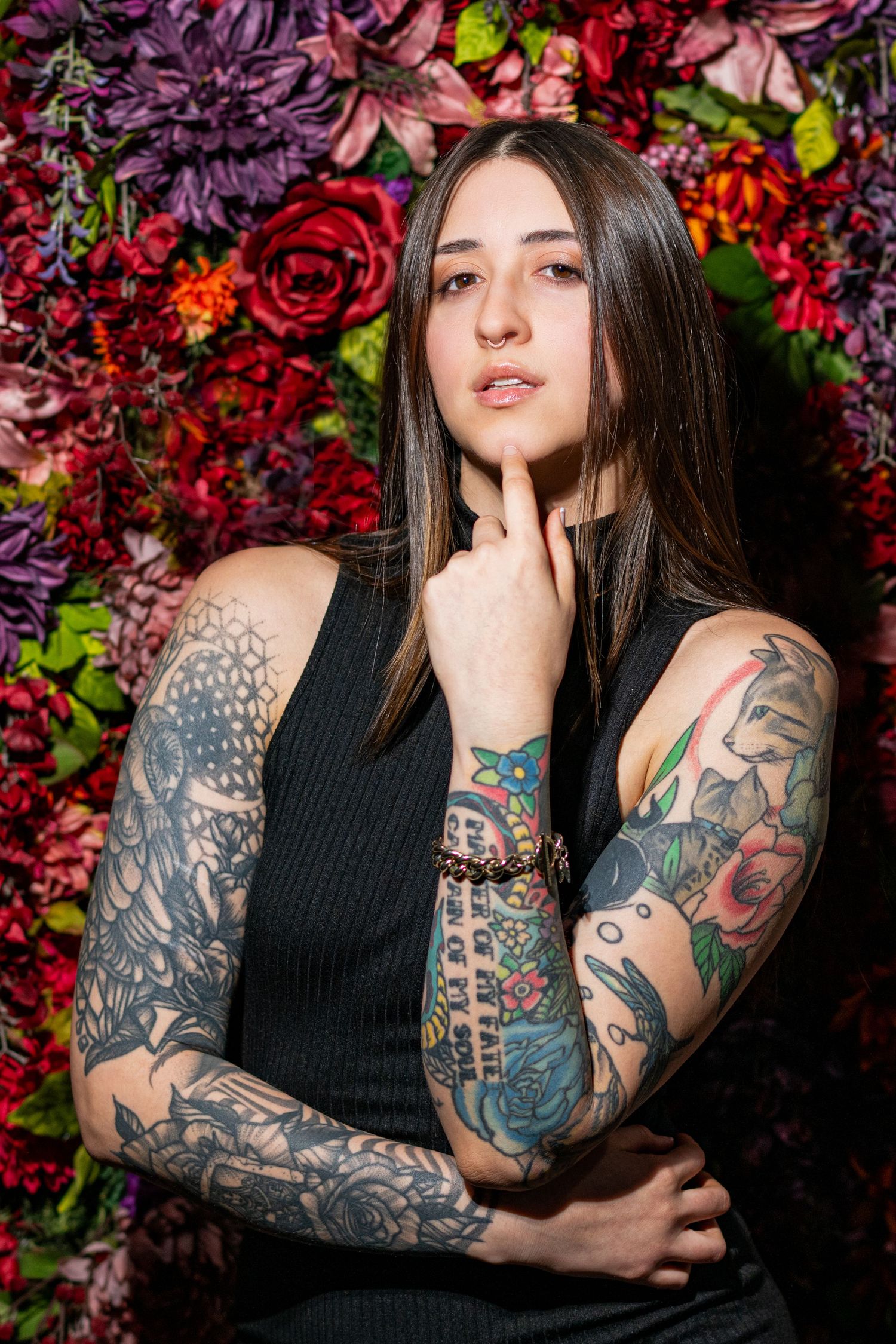 Young woman with full sleeve tattoos poses thoughtfully against a vibrant floral wall backdrop.