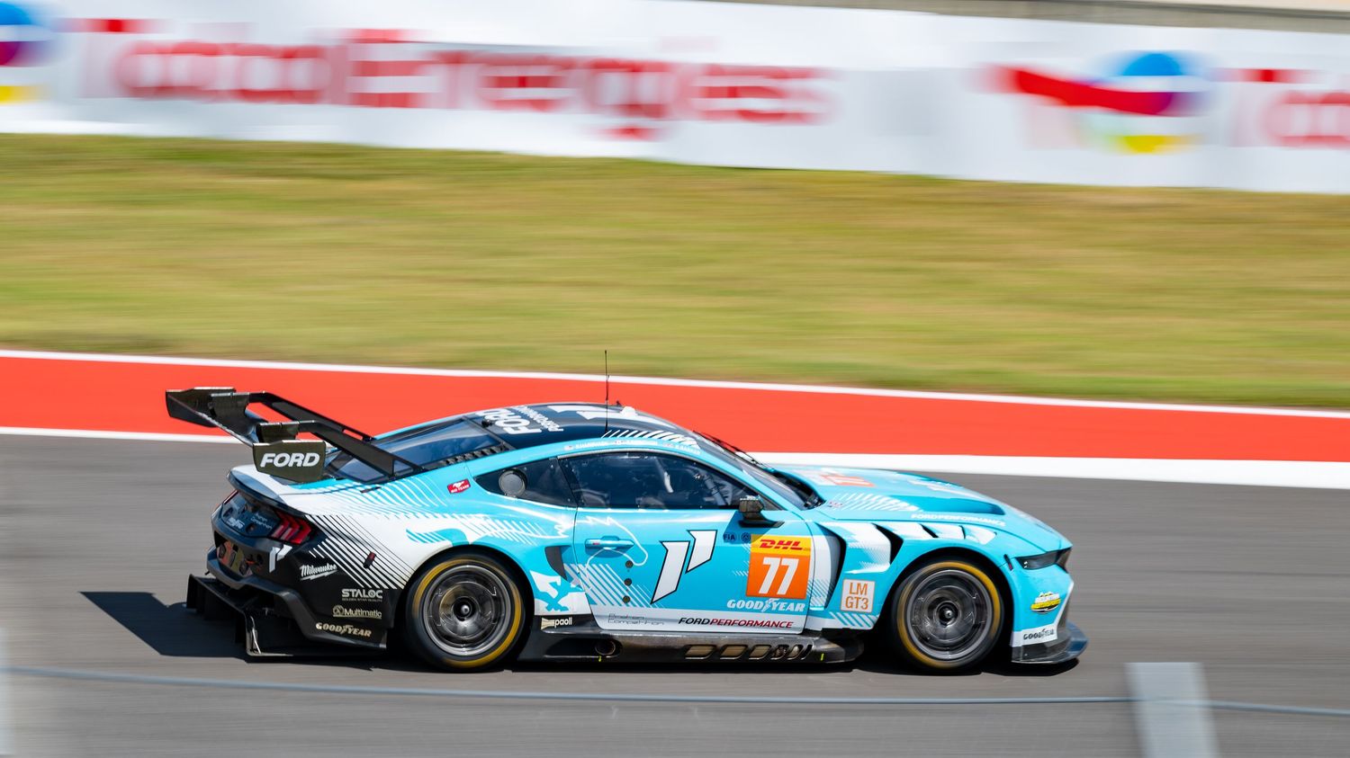 Sports cars compete on a racing circuit with colorful barriers and grass visible.