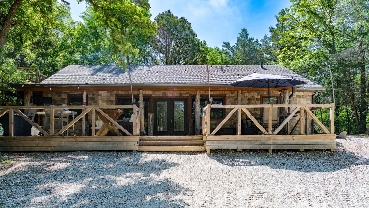 Rustic log cabin home with wooden deck and French doors surrounded by lush trees on a gravel driveway.