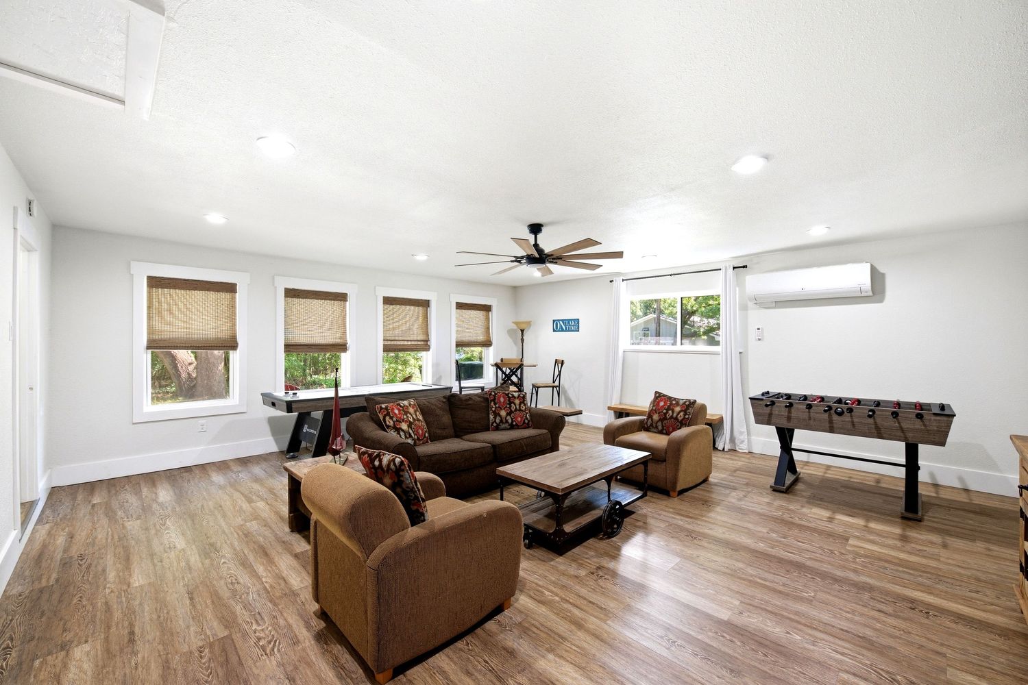 Bright living room with hardwood floors, brown sectional sofa, ceiling fan, and foosball table near a mini-split AC unit.