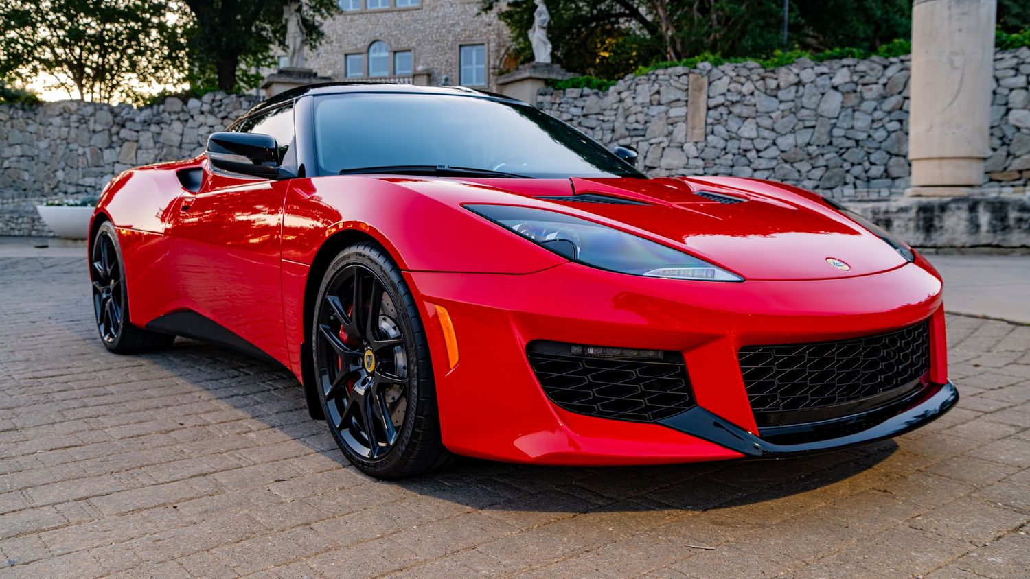 Red Lotus Evora sports car parked on cobblestone driveway with black alloy wheels and sleek convertible design.