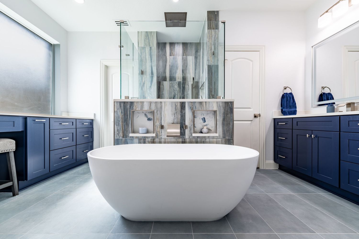 Elegant master bathroom with freestanding white soaking tub, navy blue cabinets, gray tile floors, and glass shower enclosure.