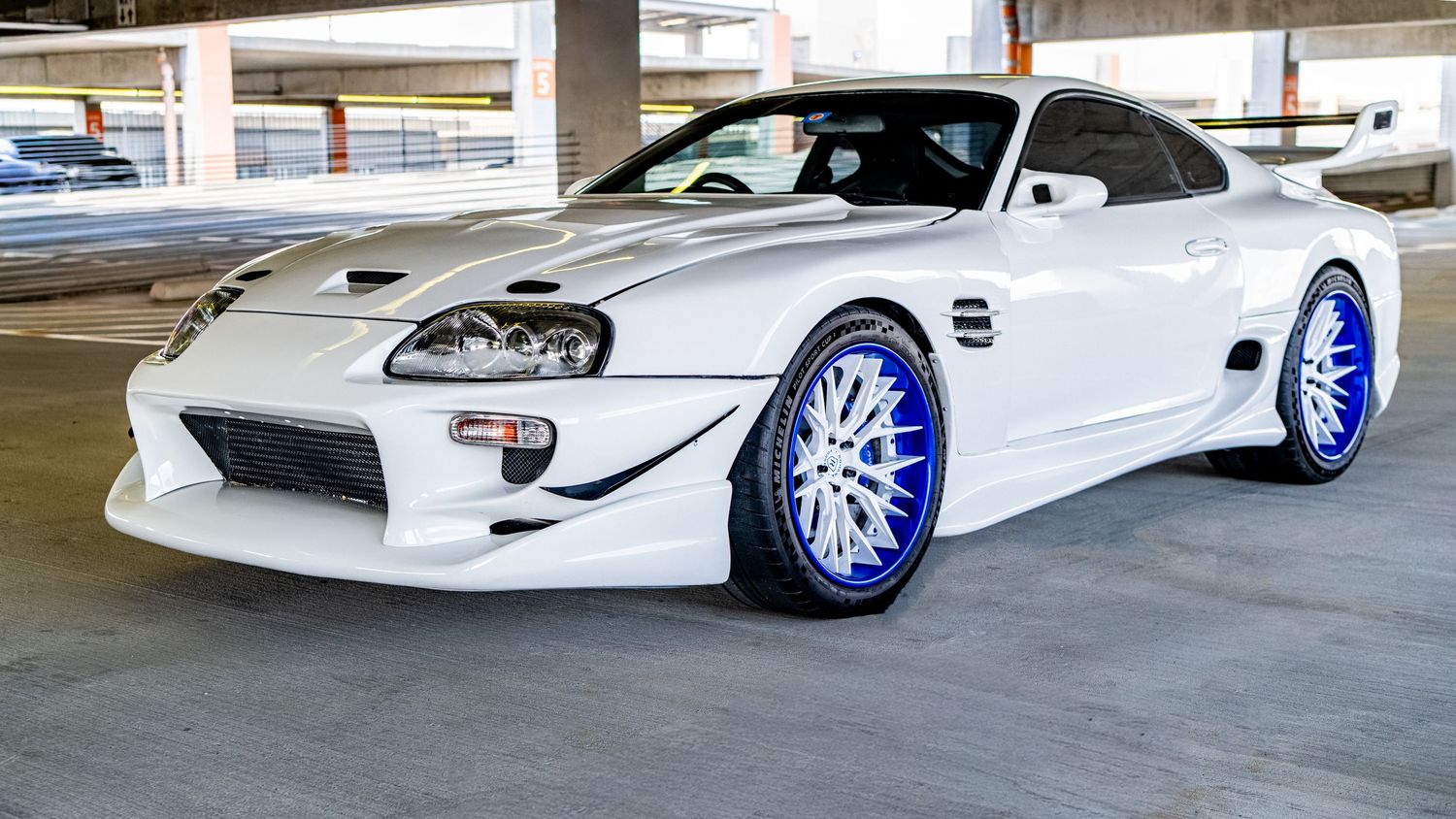 White Toyota Supra with blue chrome wheels and wide body kit parked in a garage.