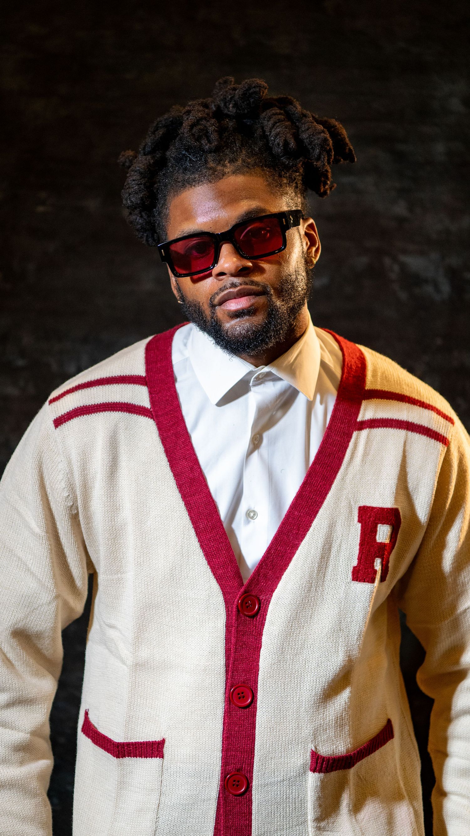Stylish portrait in cream varsity cardigan with red trim wearing sunglasses against dark background.