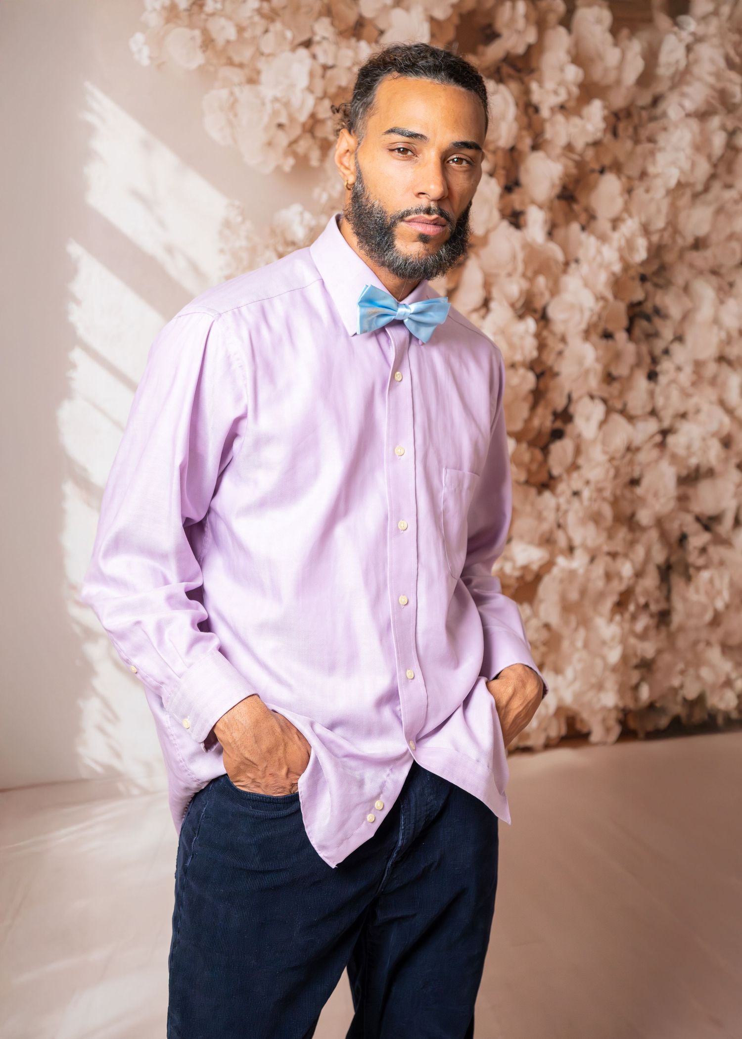 Stylish portrait of person in light purple dress shirt and blue bowtie against floral backdrop.