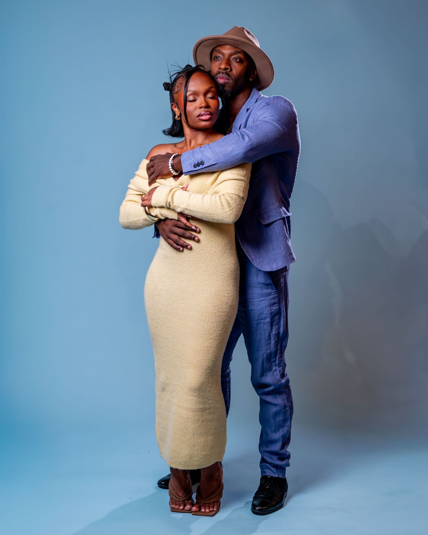 A couple embraces tenderly in a studio, woman in cream dress, man in blue blazer and hat, against a light blue background.