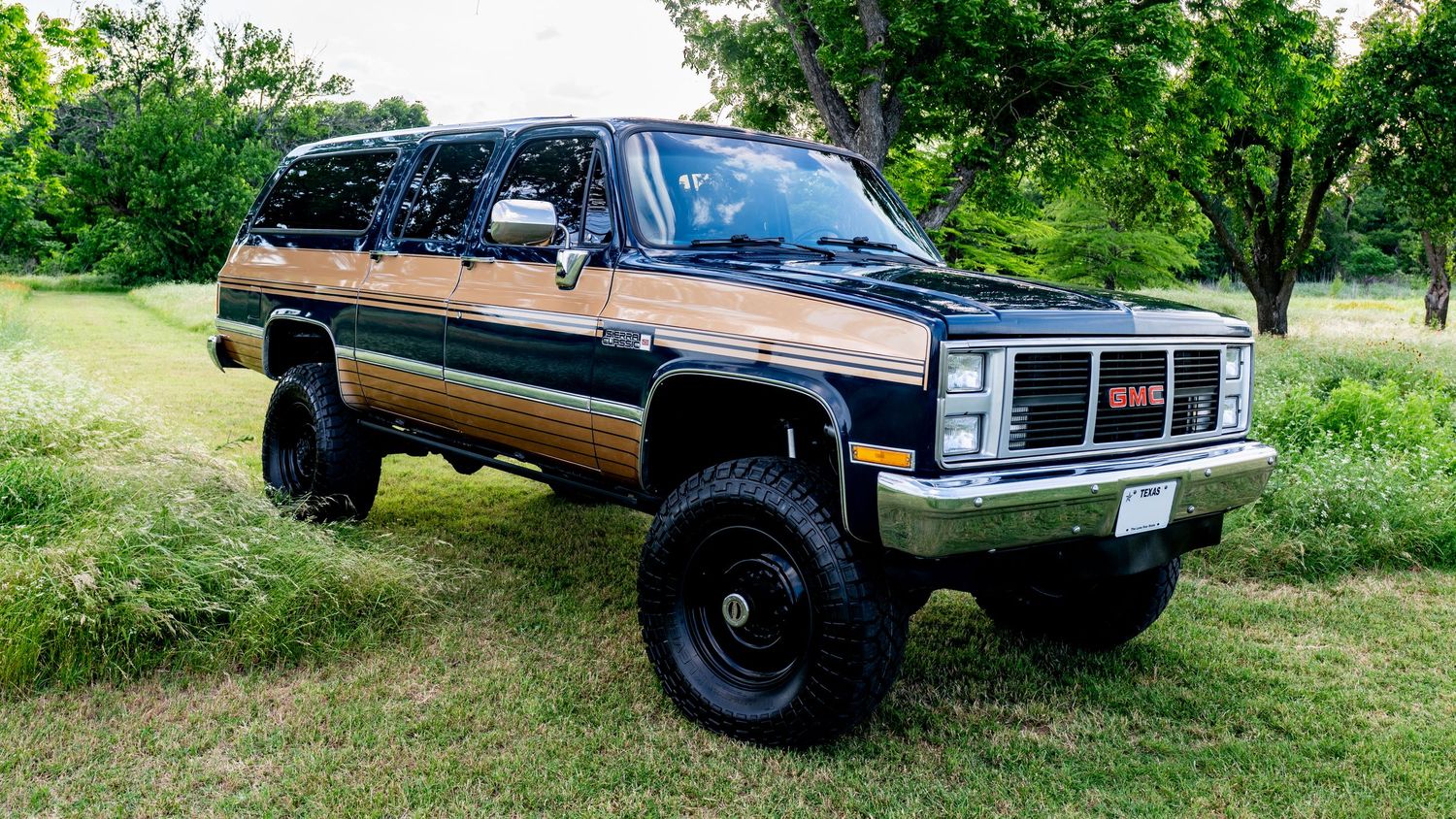 Lifted GMC Suburban with wood panel trim and black exterior parked on grass, featuring oversized off-road tires.