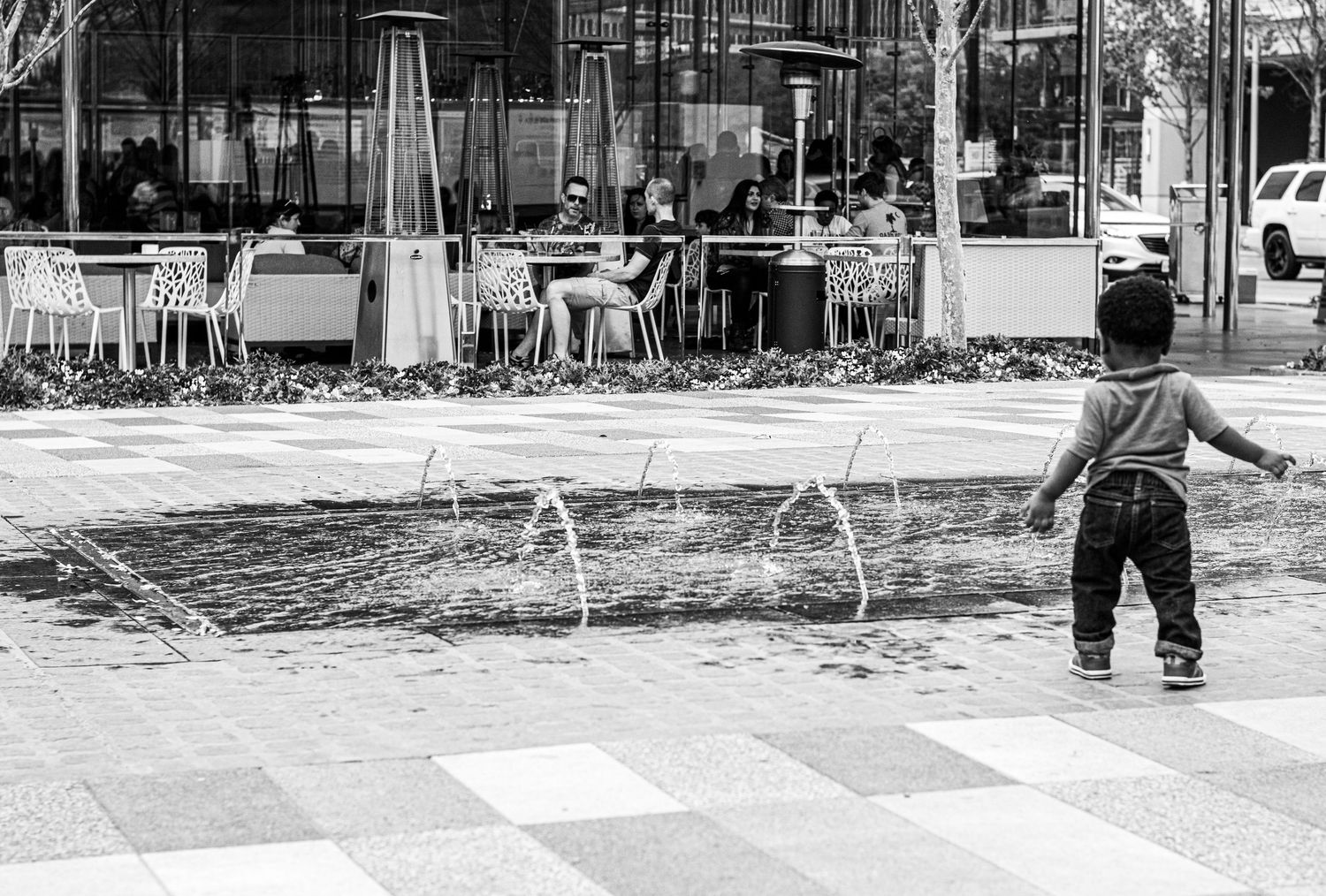 A small child watches water fountains spray up from the ground near an outdoor cafe terrace.