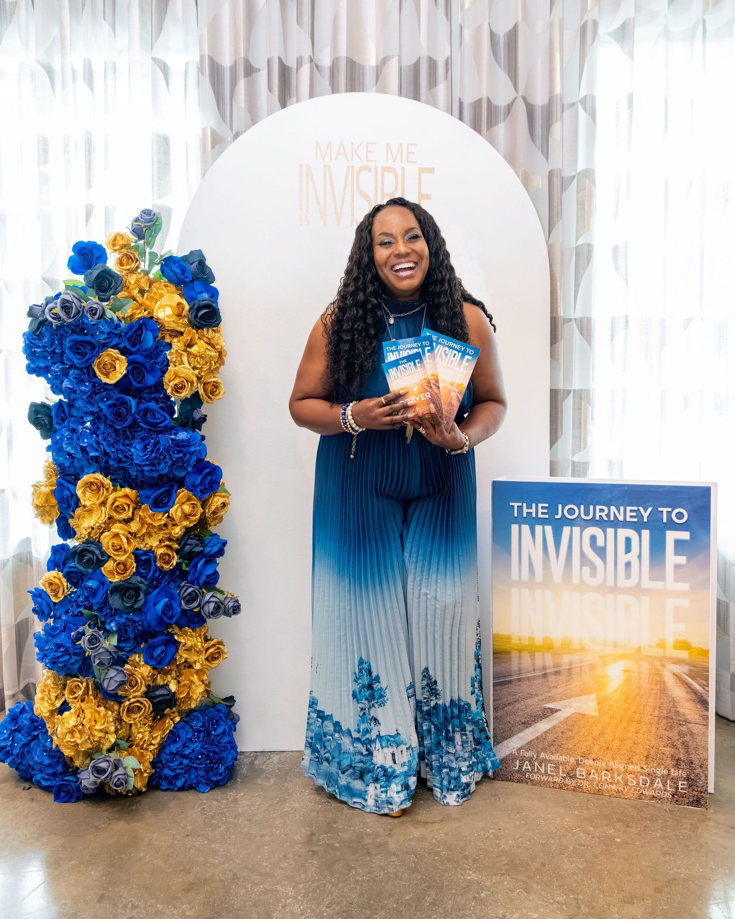 Woman in blue floral jumpsuit holds books beside flower column at The Journey to Invisible book launch event.