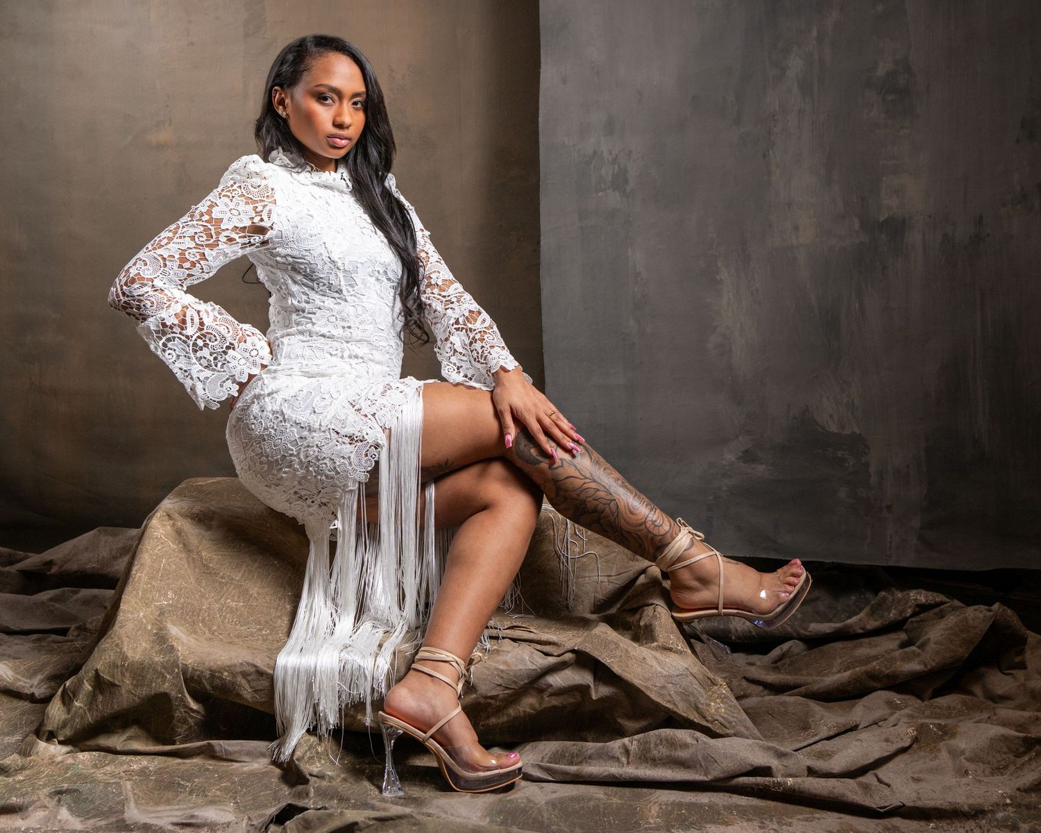 Woman in white lace mini dress and strappy heels poses elegantly on draped fabric in a studio setting.