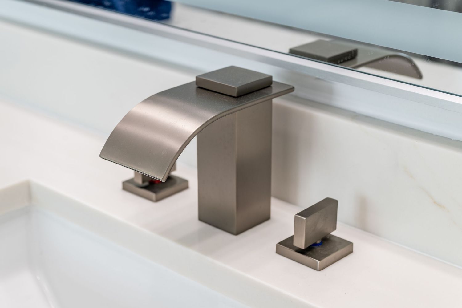 Brushed nickel widespread bathroom faucet with waterfall spout and square handles on white countertop.