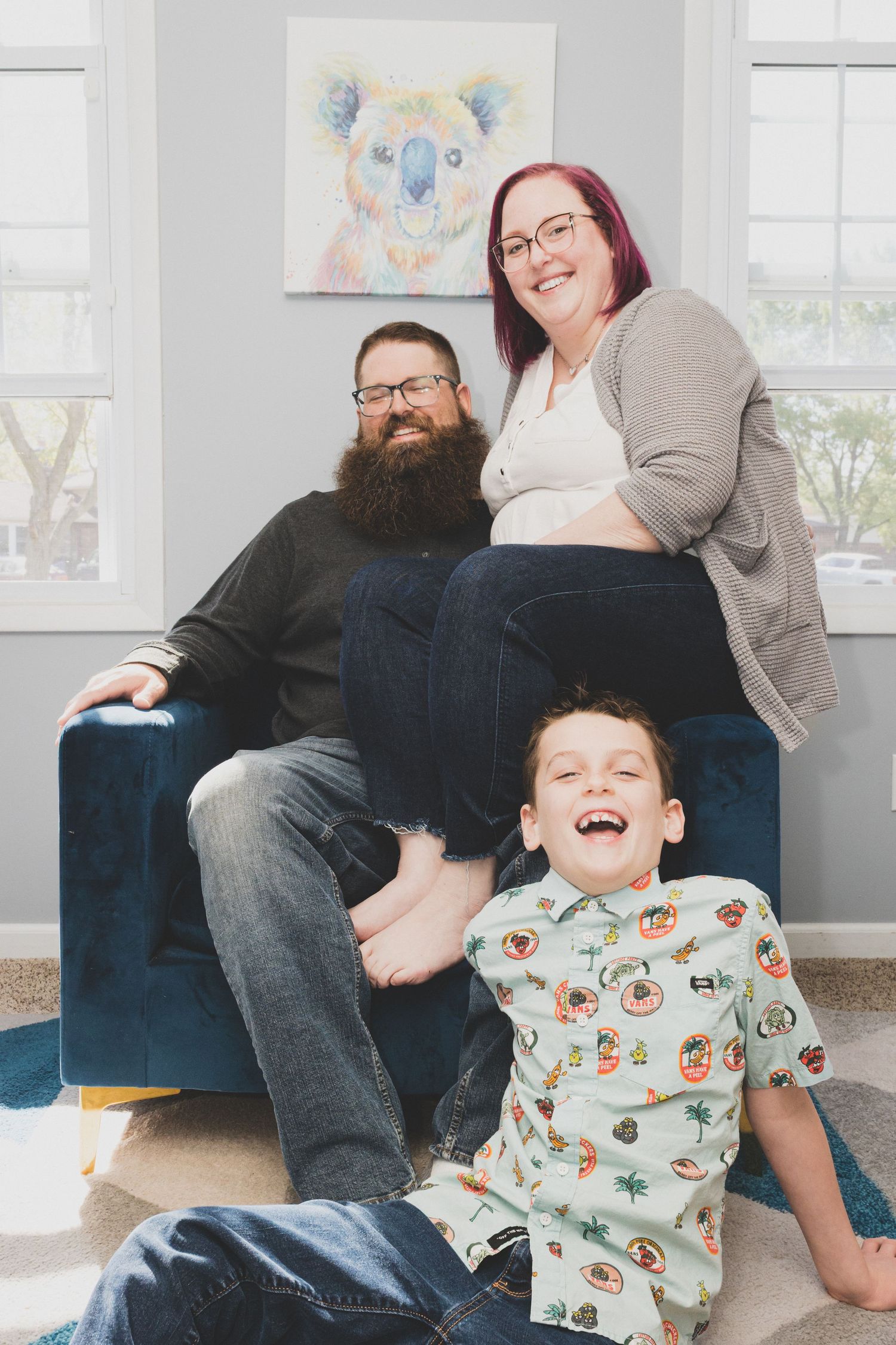 Happy family portrait with bearded dad, pregnant mom, and laughing young boy sitting together in a cozy living room.