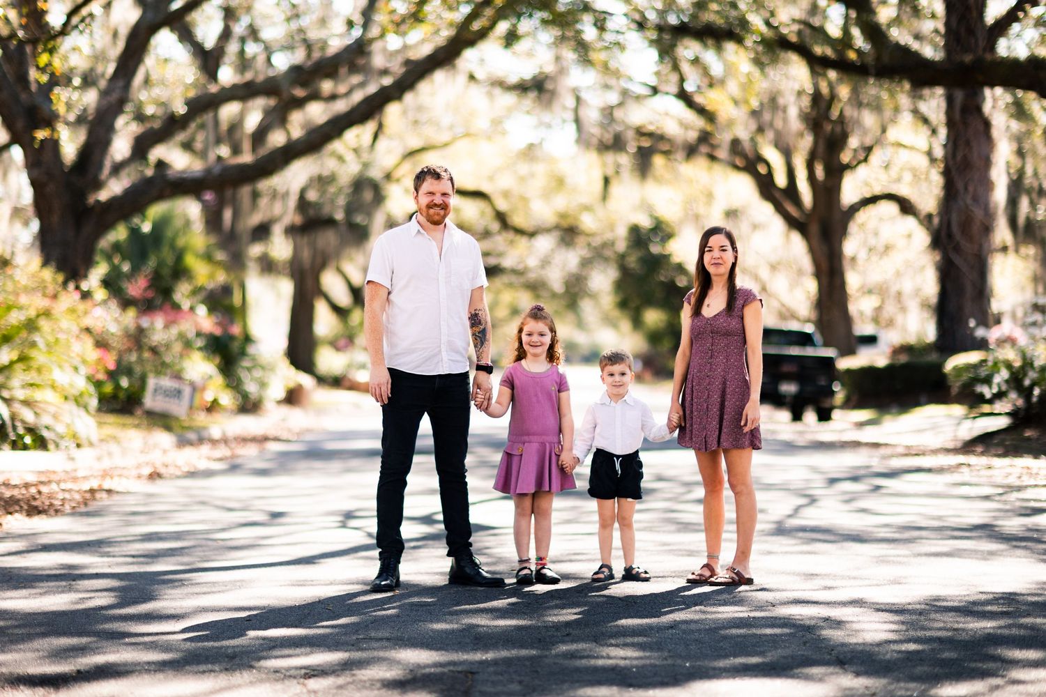 Family of four poses on a tree-lined street, parents holding hands with two young children in coordinated outfits.
