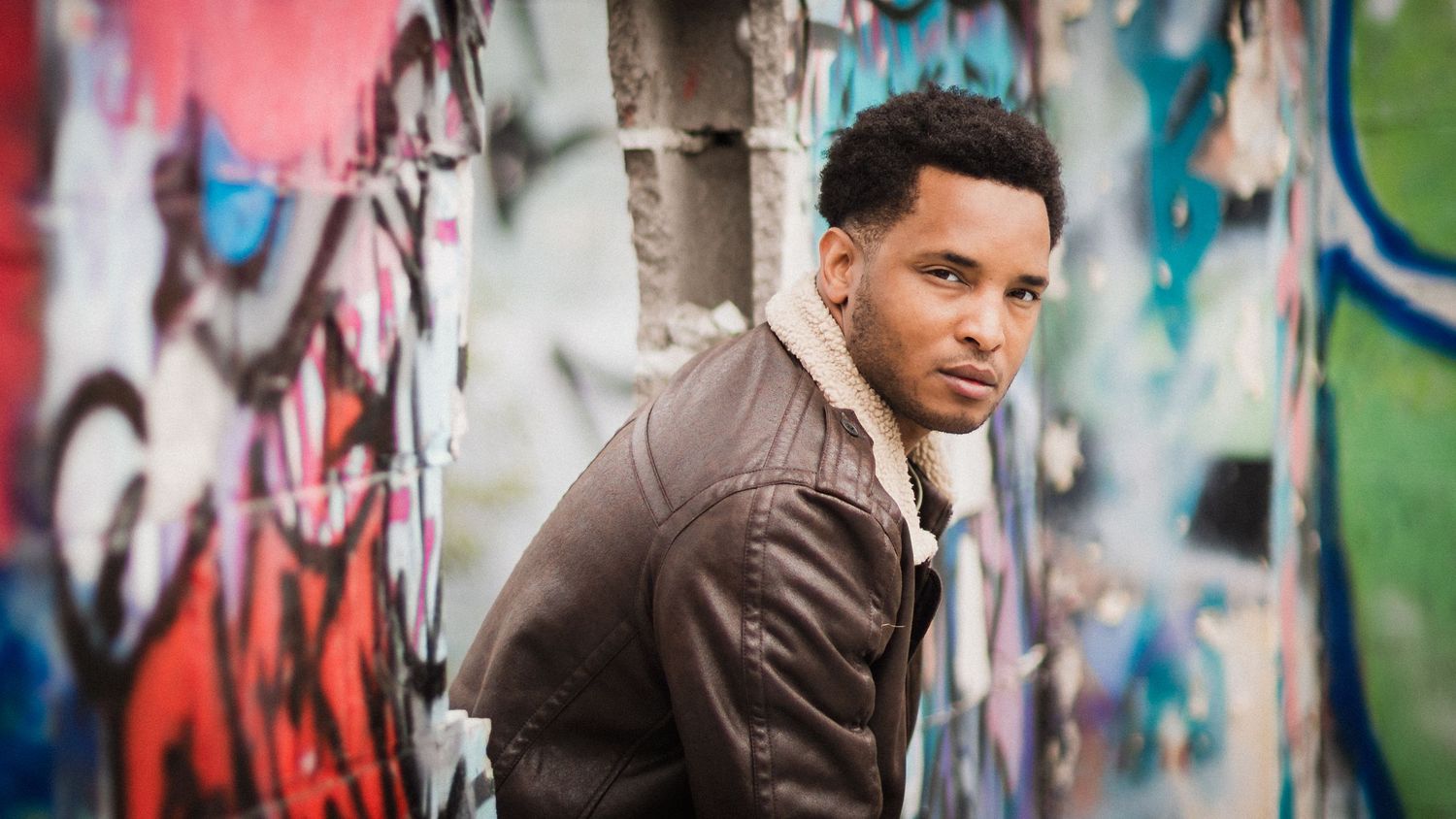 Young man in brown leather jacket posing against colorful graffiti wall in urban outdoor setting.
