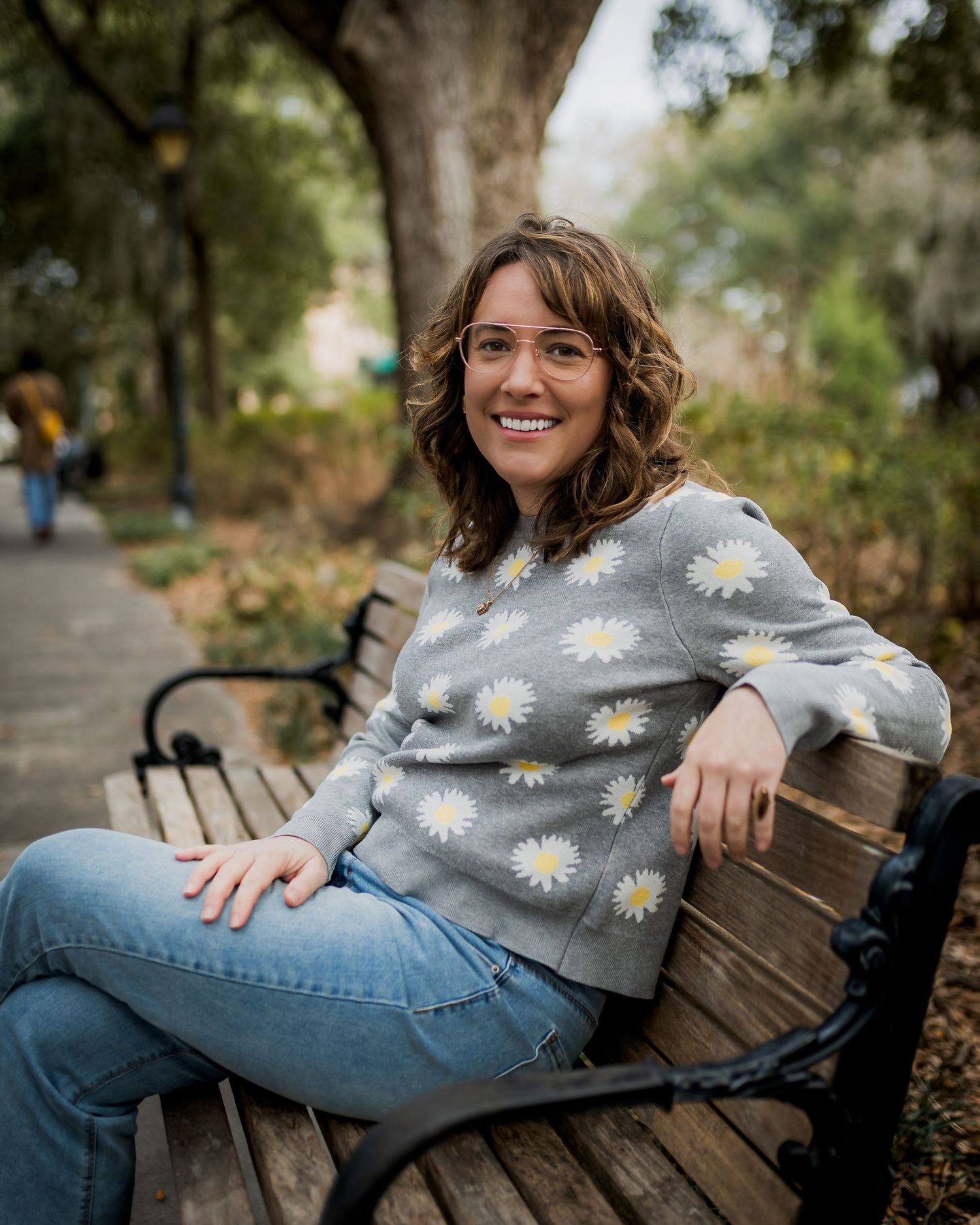 Smiling woman in daisy sweater and jeans sitting on park bench surrounded by trees.