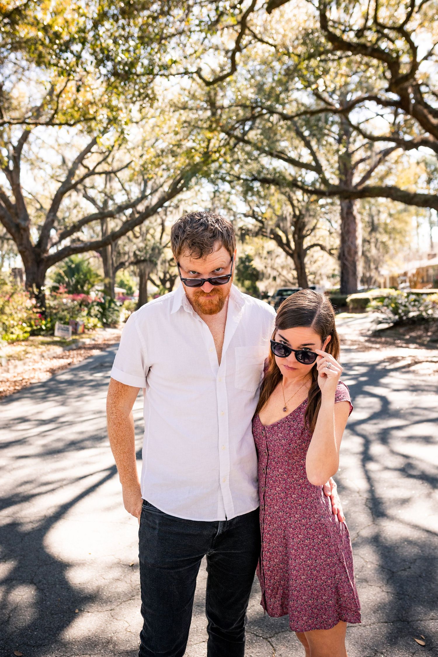 A couple in sunglasses poses on a sunny tree-lined street, woman in floral dress, man in white shirt.