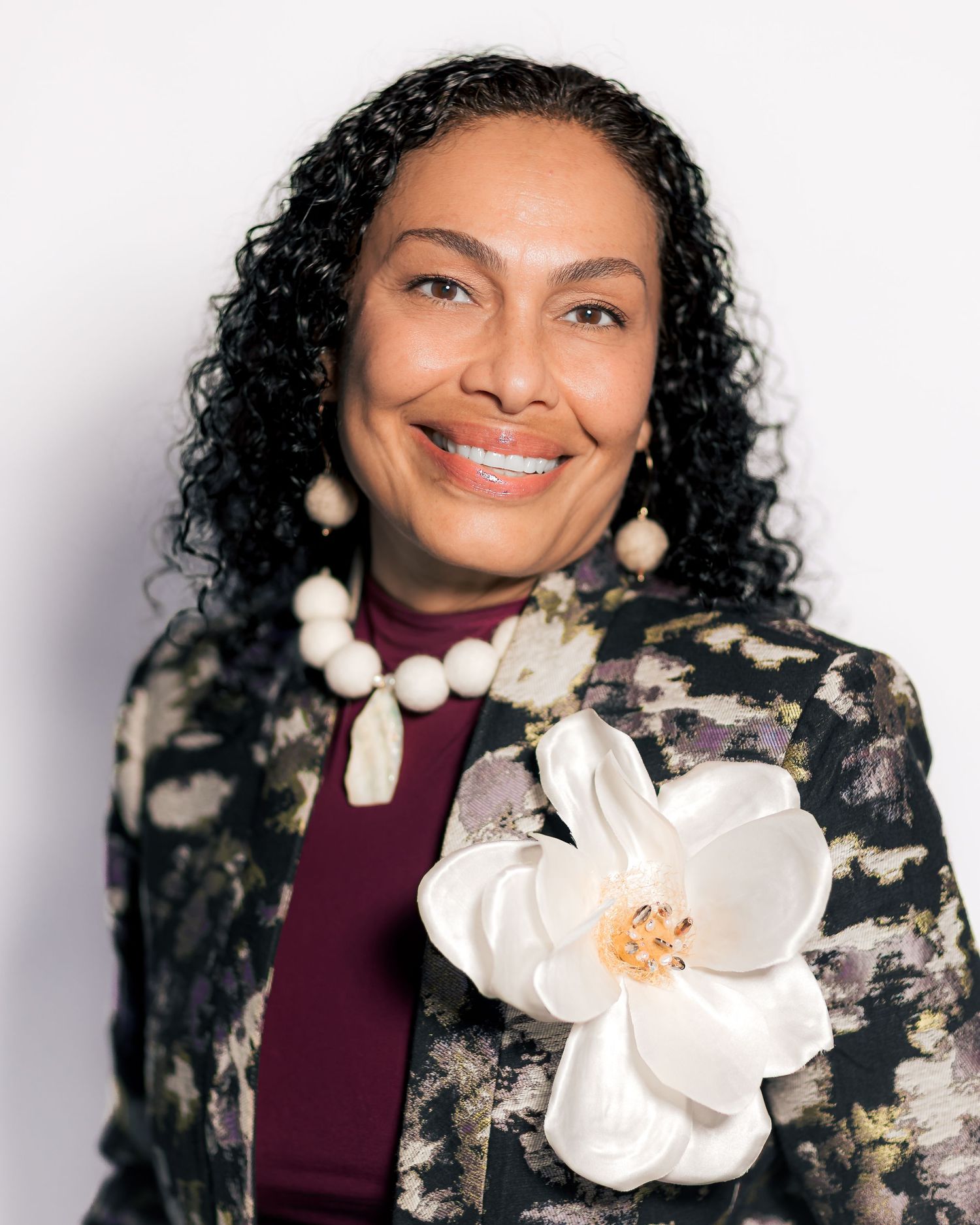 Professional woman smiling in floral blazer with white magnolia brooch, beaded necklace, and curly hair against white background.