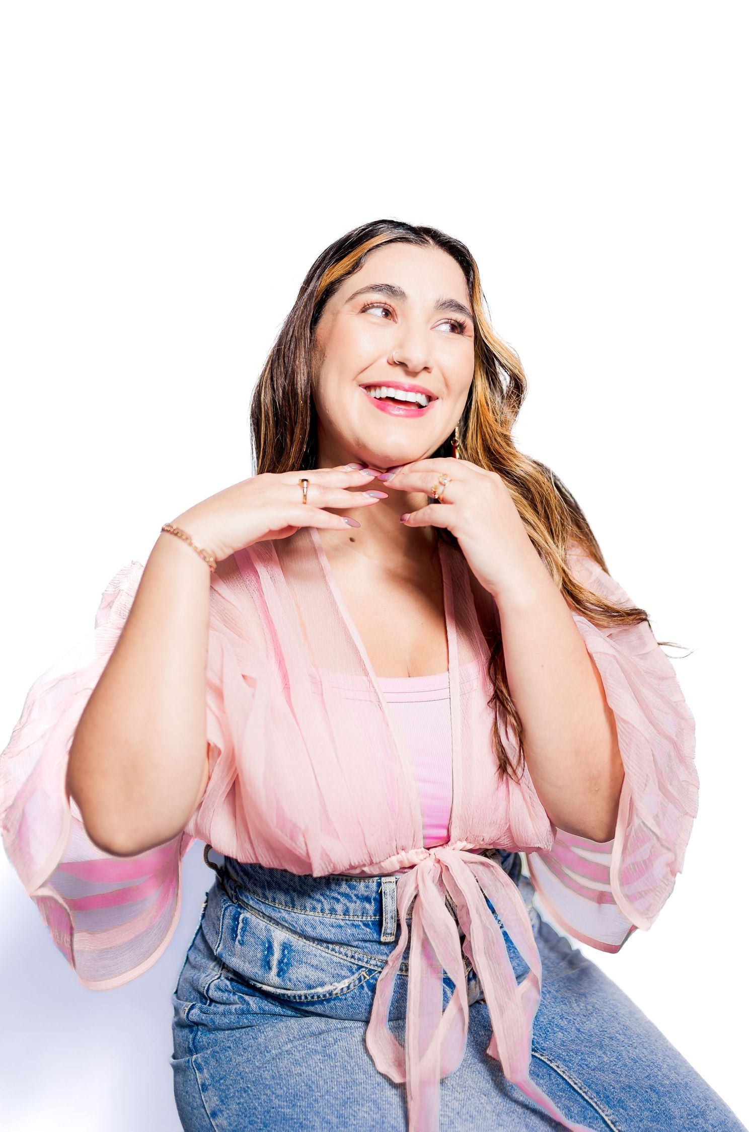 Smiling woman in pink sheer blouse and jeans posing with hands near chin against white background.