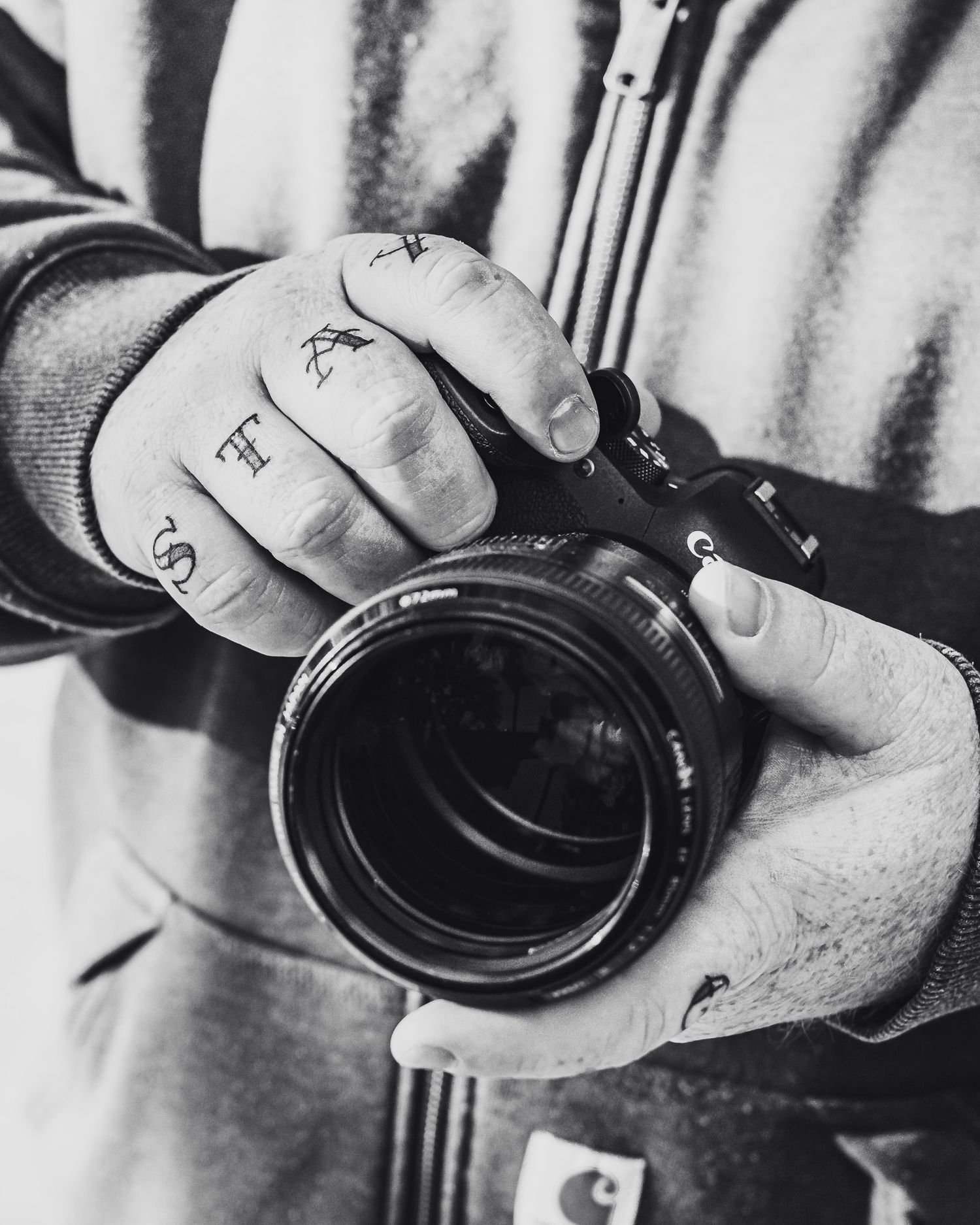 Black and white photo of tattooed hands holding a DSLR camera with a large lens, wearing a Carhartt jacket.