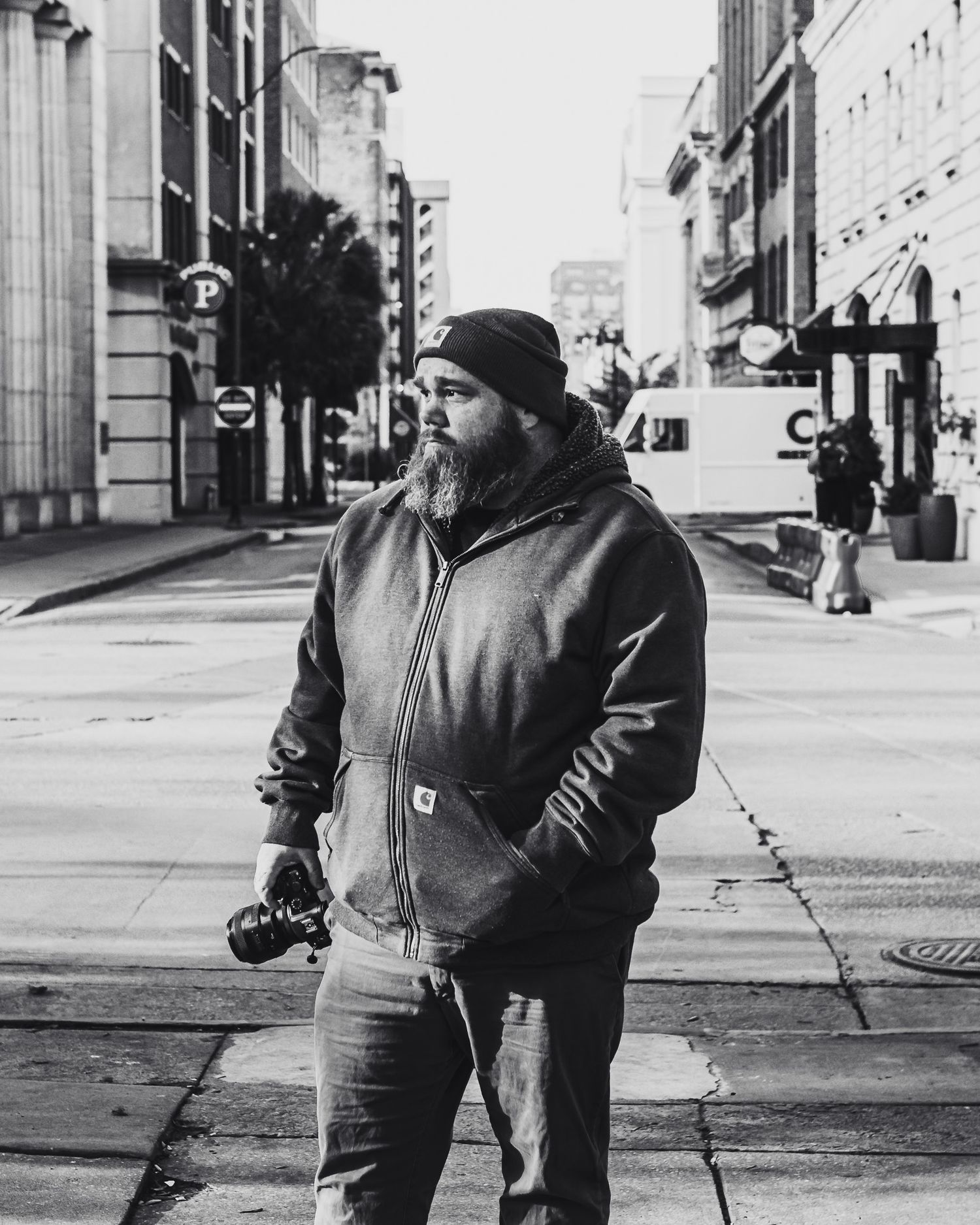 Bearded man in Carhartt hoodie and beanie holds camera on urban street in black and white photography.