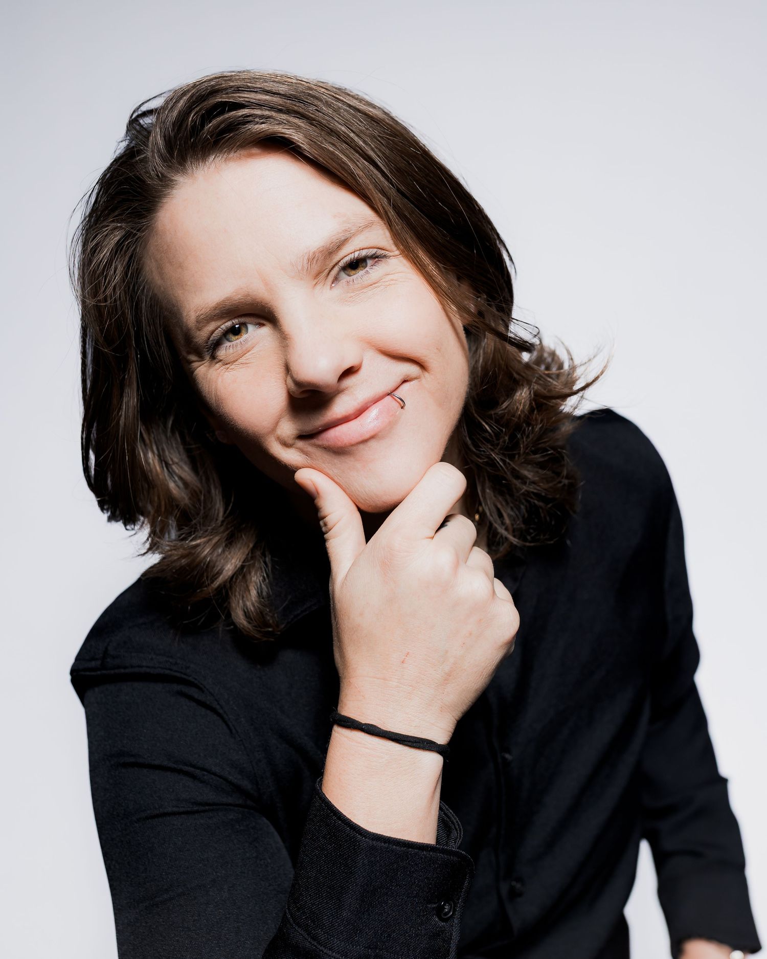 Woman with dark hair in black jacket smiling confidently, resting chin on hand against white background.
