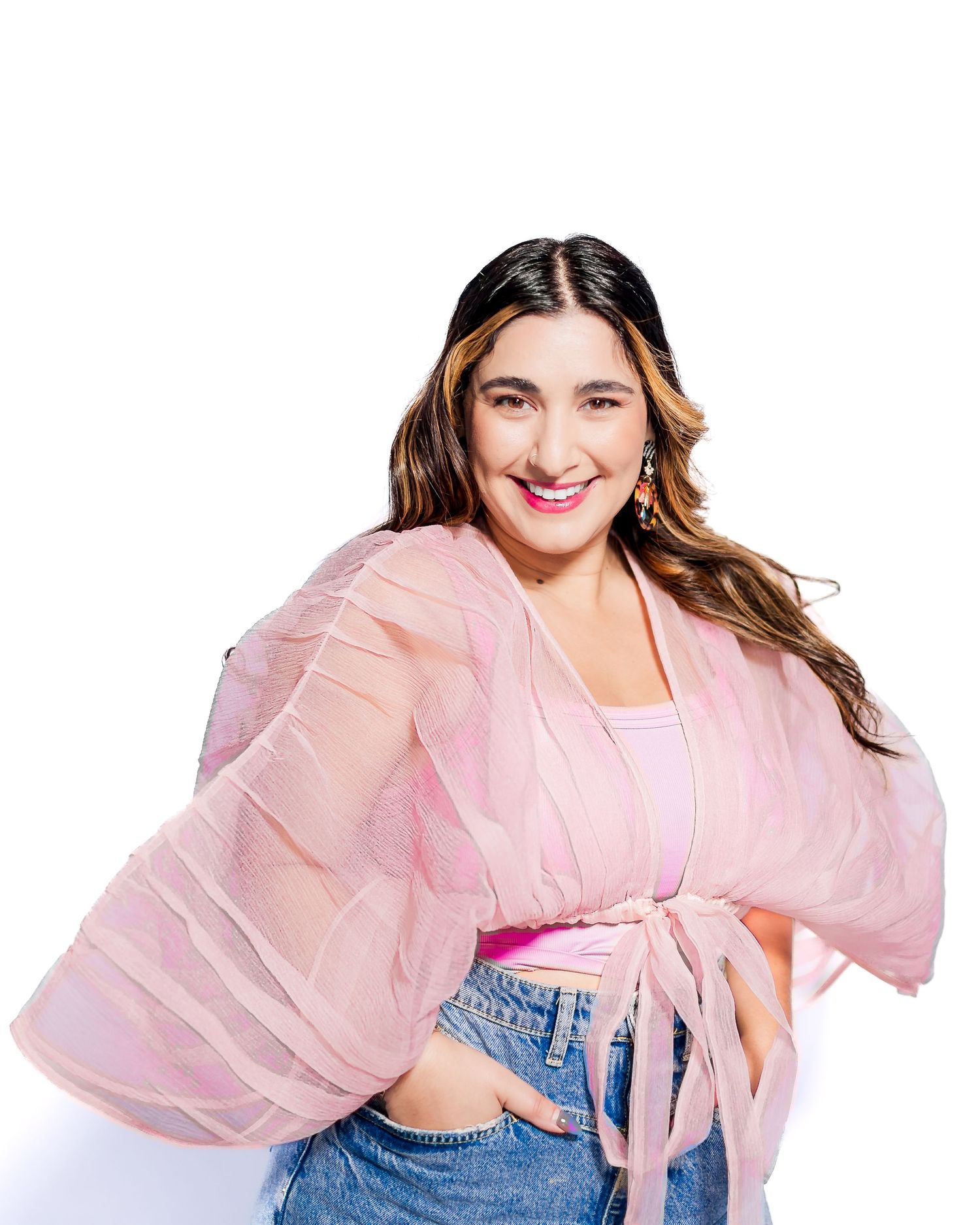 Woman in pink sheer kimono top and jeans smiling against white background.