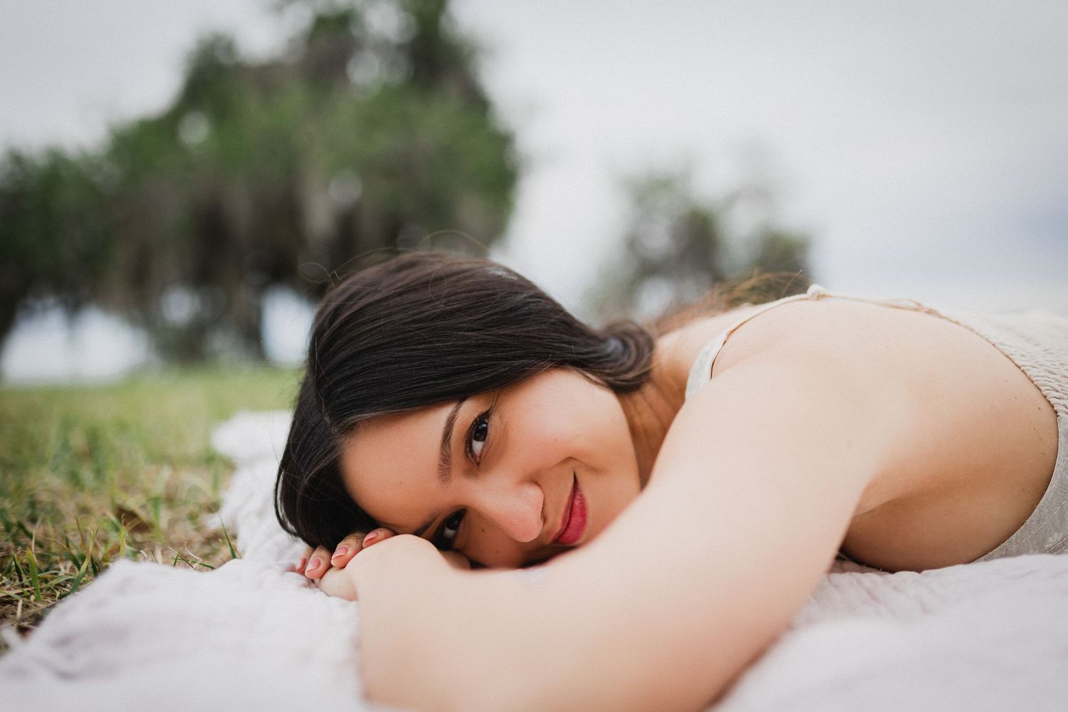 Young woman with dark hair smiling while lying on a white blanket outdoors near green trees.